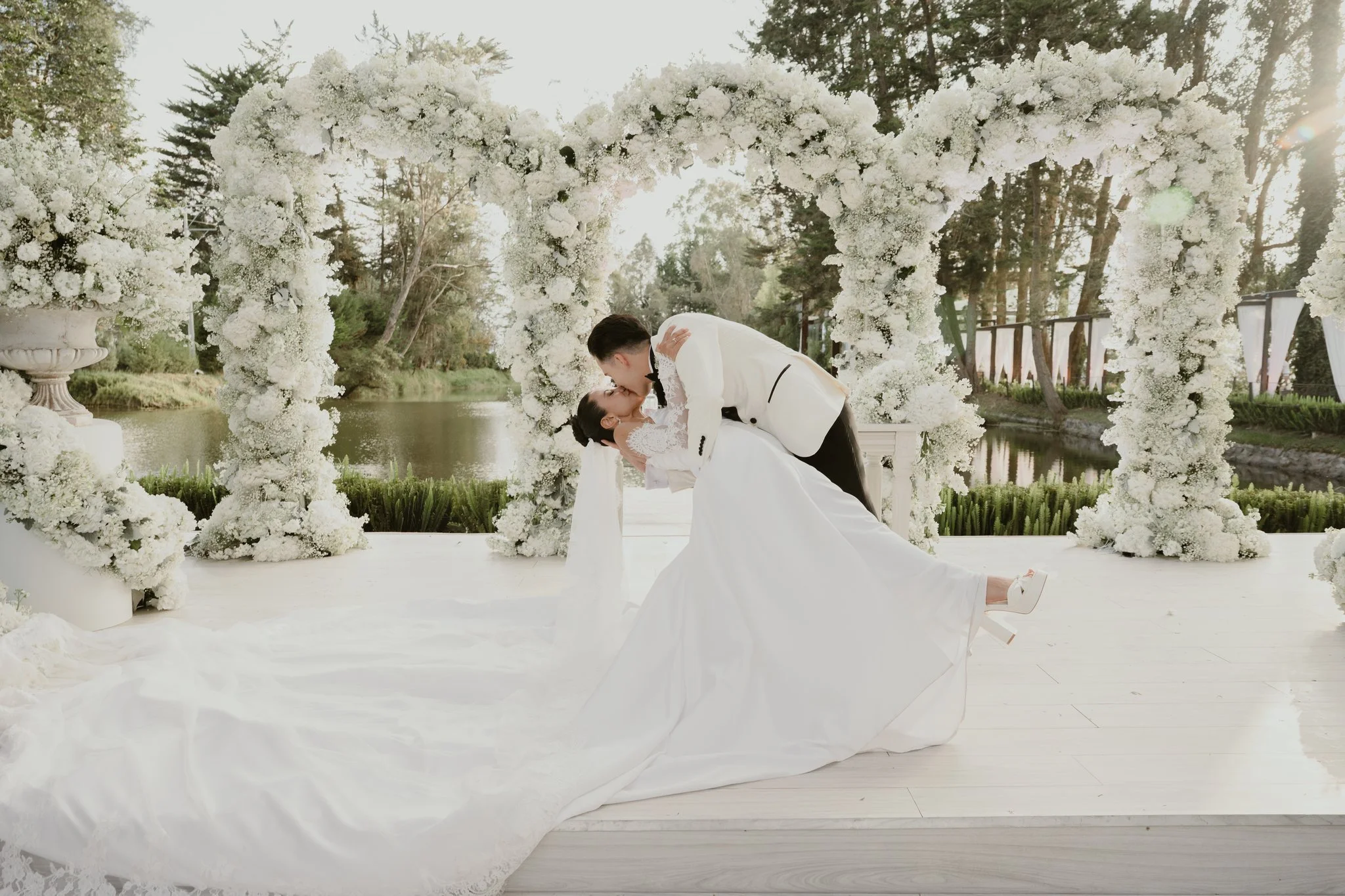 A bride and groom share a kiss outdoors during their wedding celebration, with a floral arch and trees in the background.
