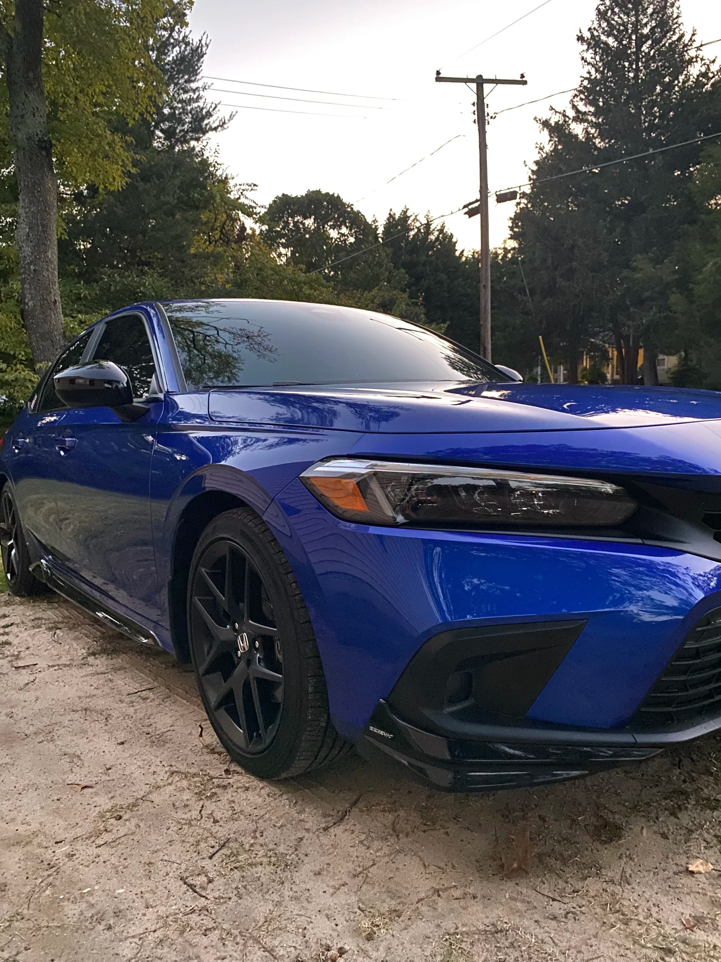 A blue sports car parked on a dirt surface with trees and power lines in the background during dusk.