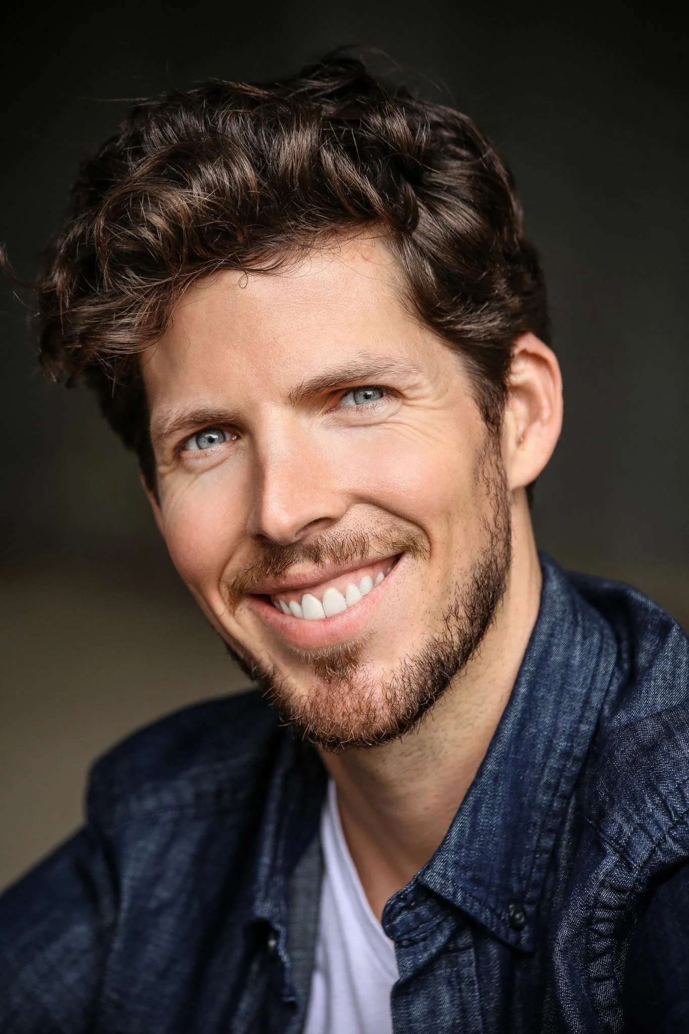 Close-up portrait of a smiling man with curly brown hair, blue eyes, and a beard, wearing a dark shirt.