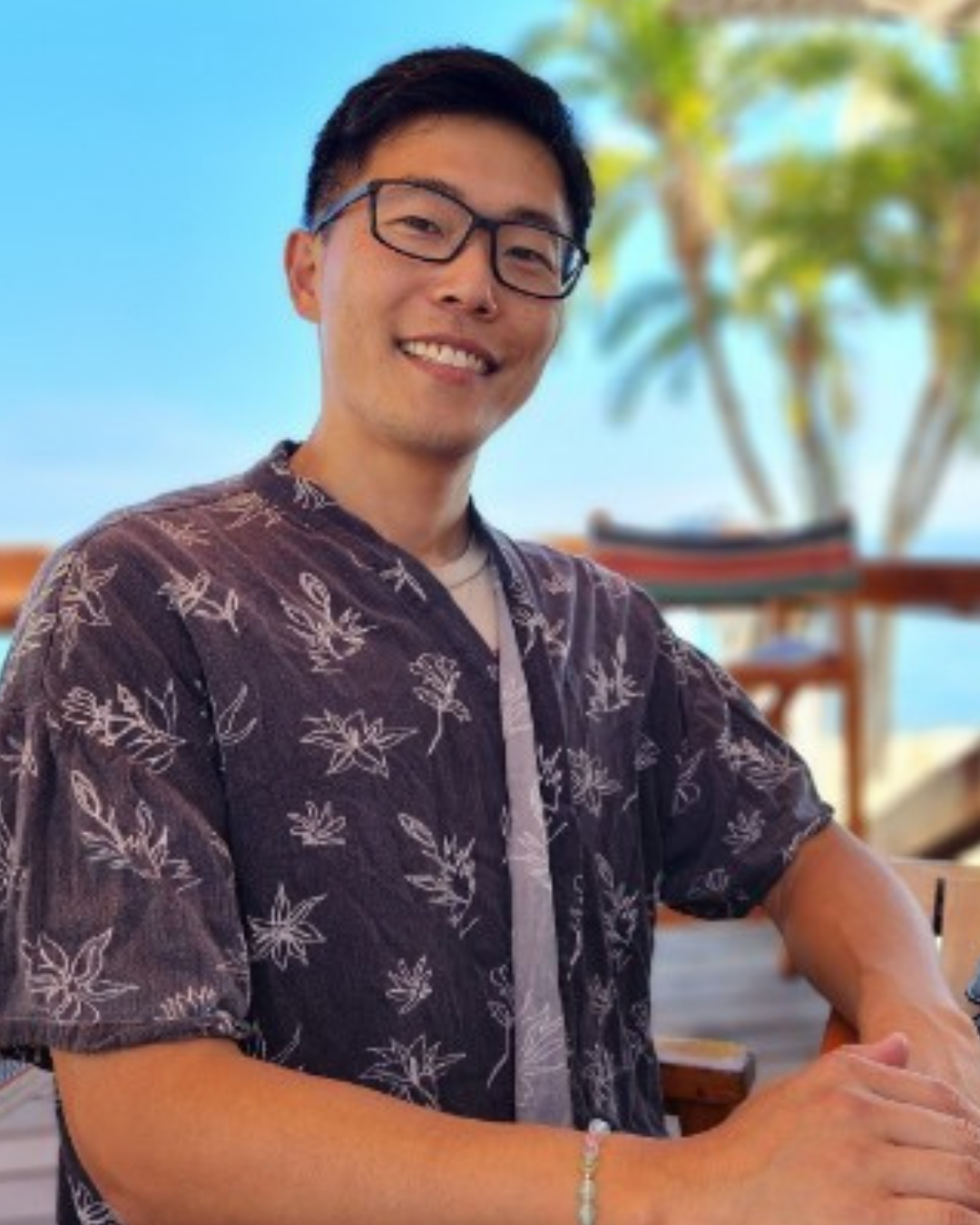 A man with black hair, glasses, and a light beard smiling outdoors during daytime. He is wearing a dark floral shirt and sitting at a table with a blurred background of trees and sky.