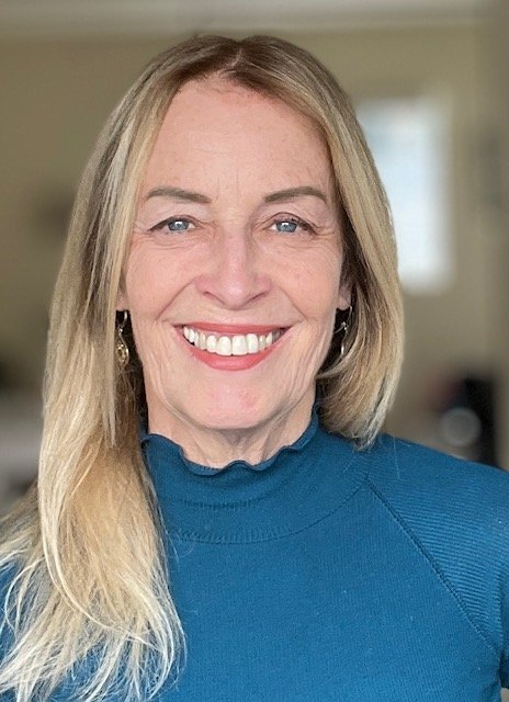 Close-up portrait of a smiling woman with blonde hair, wearing a blue turtleneck, in an indoor setting.