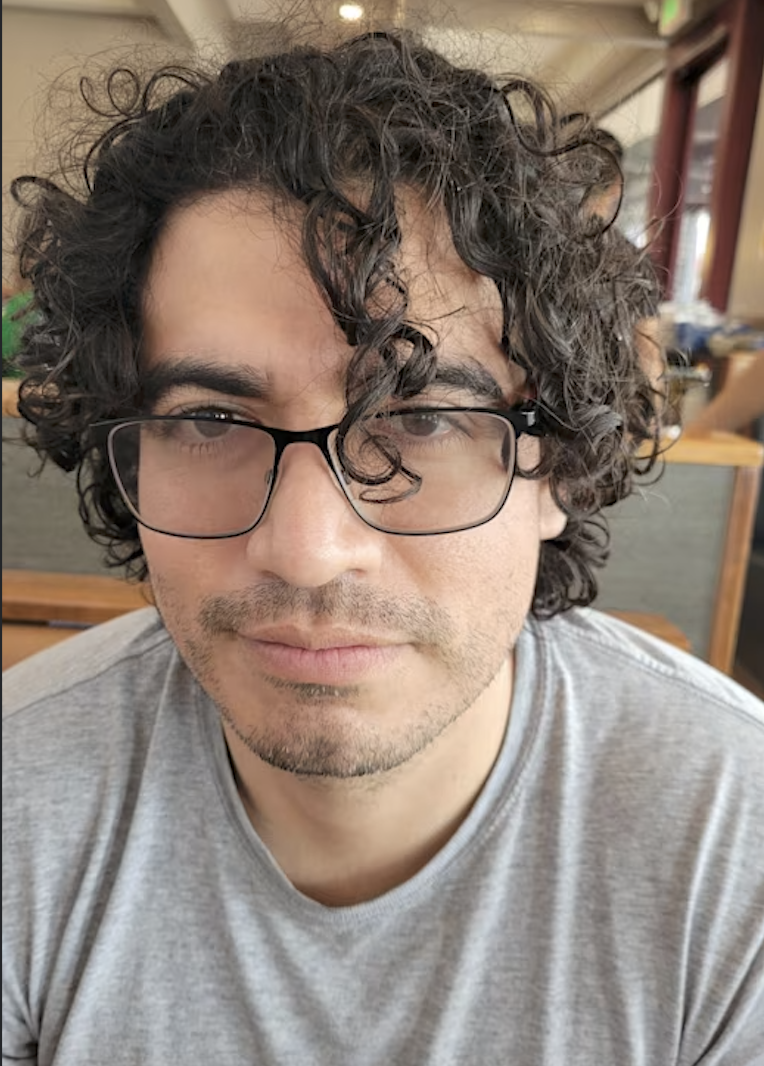 Close-up of a man with curly dark hair, glasses, and light skin, wearing a gray T-shirt, sitting indoors at a restaurant or cafe.
