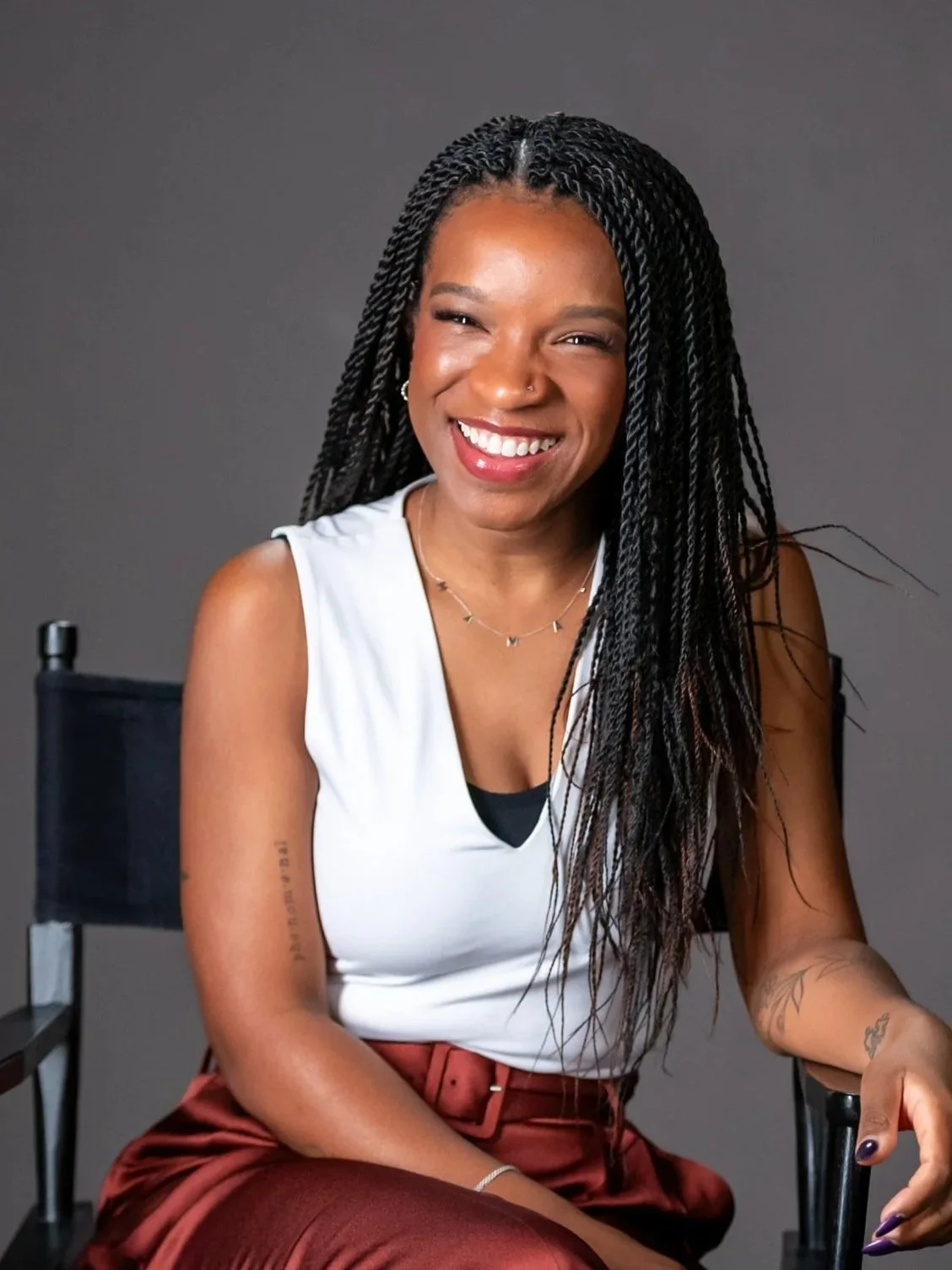 Smiling woman with long black braids wearing a white sleeveless top and maroon pants, sitting in a black chair against a gray background.