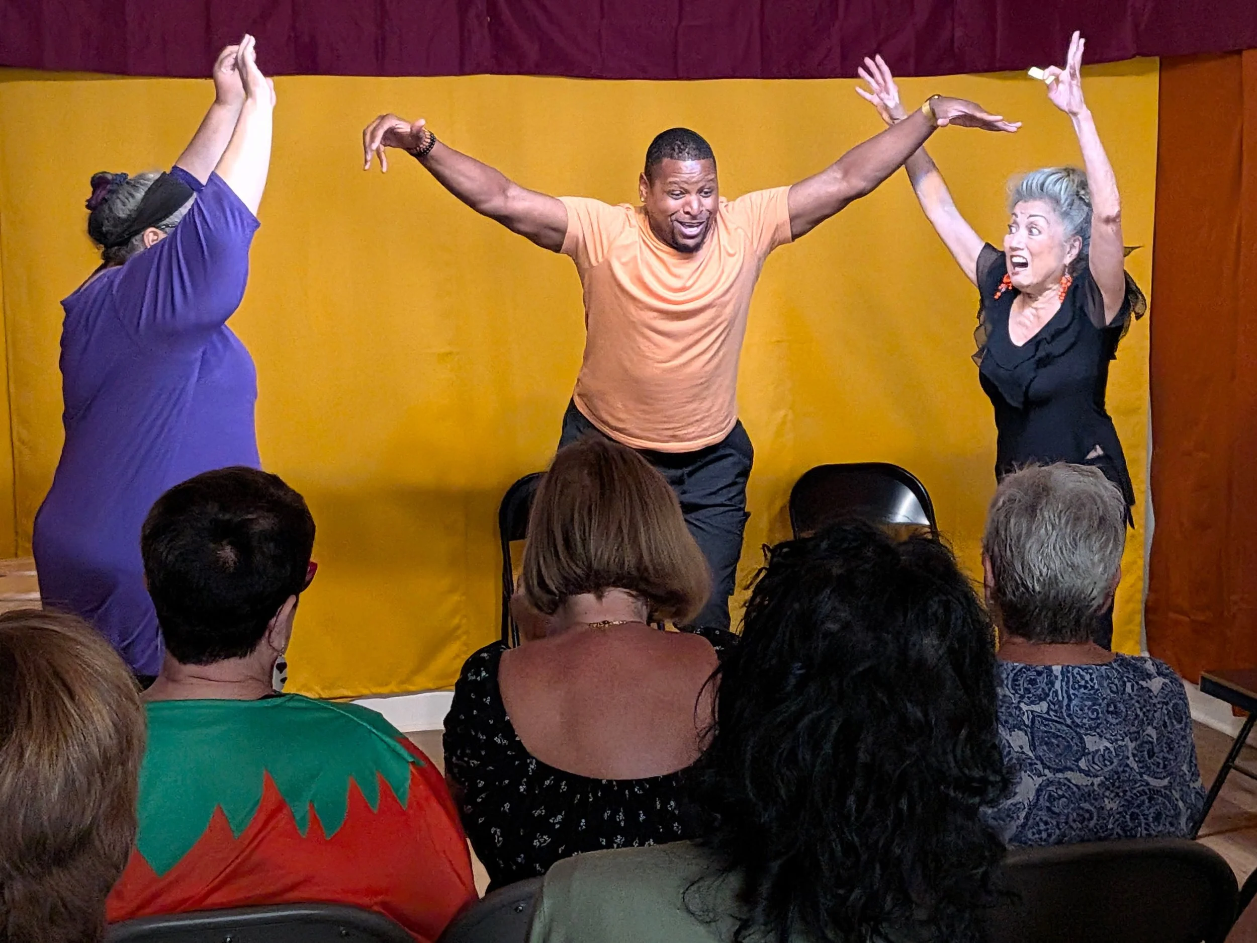 A man with short hair and wearing an orange t-shirt is dancing with his arms raised. Two women on either side of him are dancing with their arms raised, one in a purple top and the other in a black top with orange earrings. The scene appears to be a lively performance or dance event with an audience seated in front.