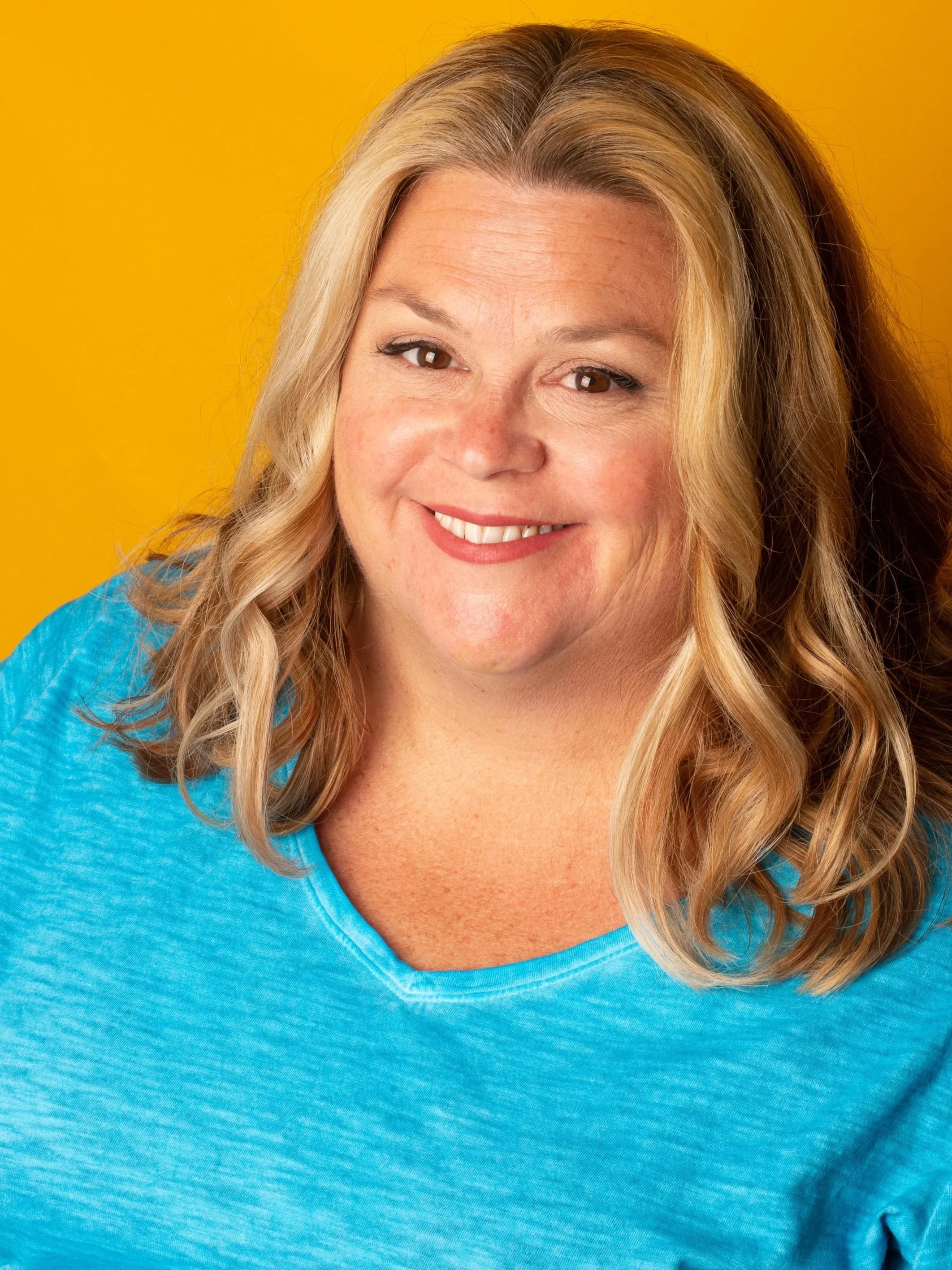 Close-up of a smiling woman with long blonde hair, wearing a bright blue shirt, against a yellow background.