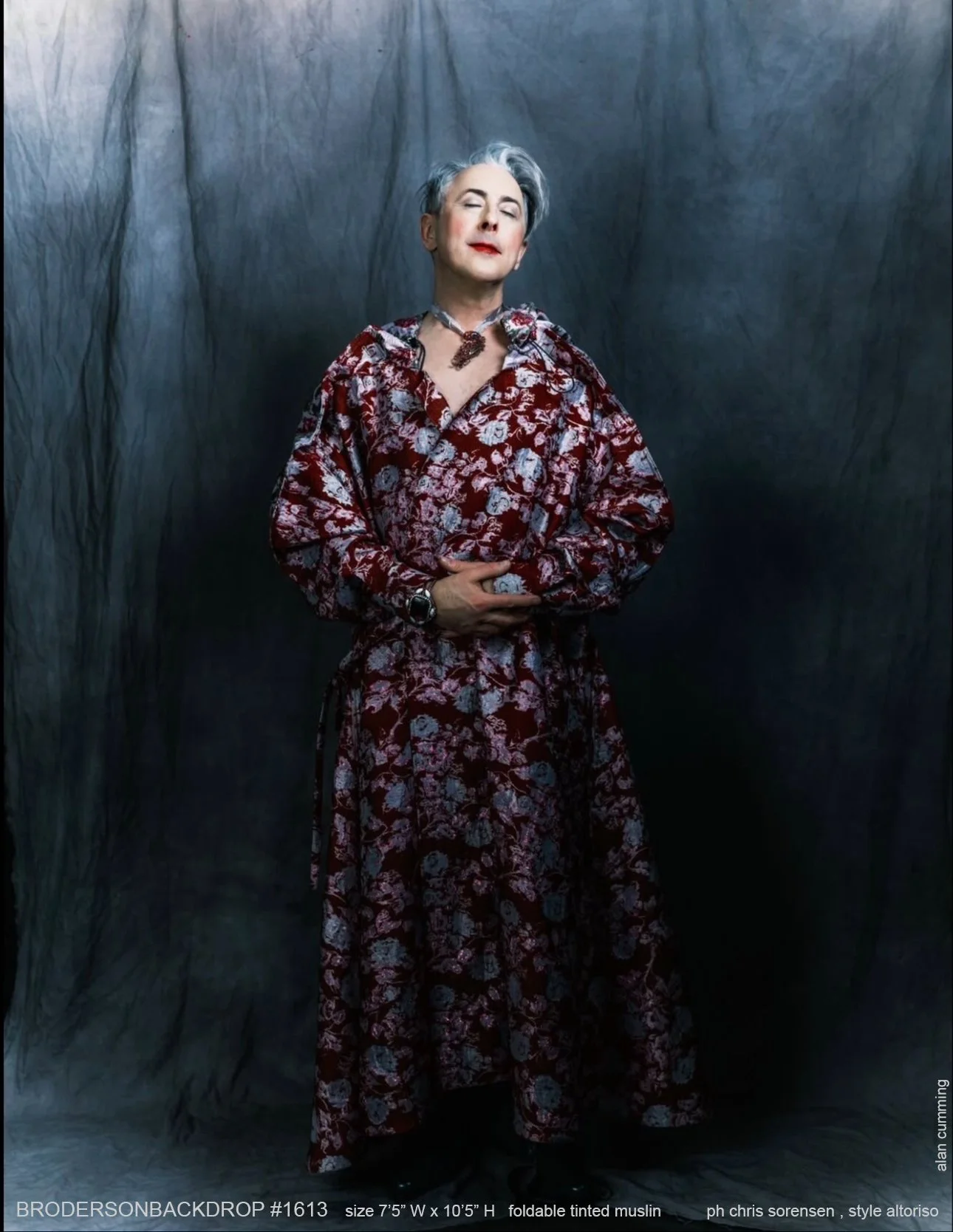 A person with short, gray hair and fair skin posing with eyes closed, wearing a long, floral red and white dress, a necklace with a large pendant, and a watch, standing against a dark background.