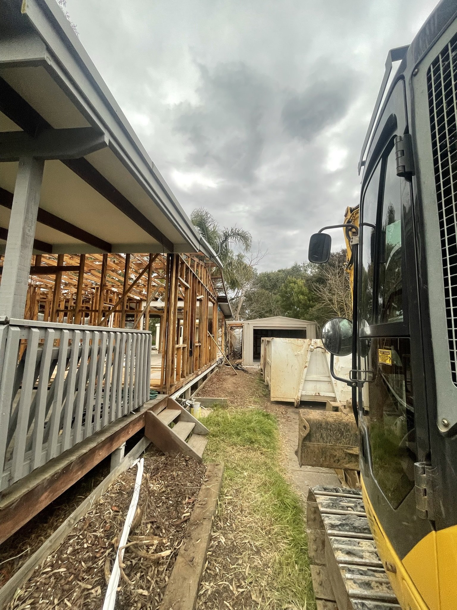 Construction site with partially built wooden structure, excavator, and containers, under cloudy sky.