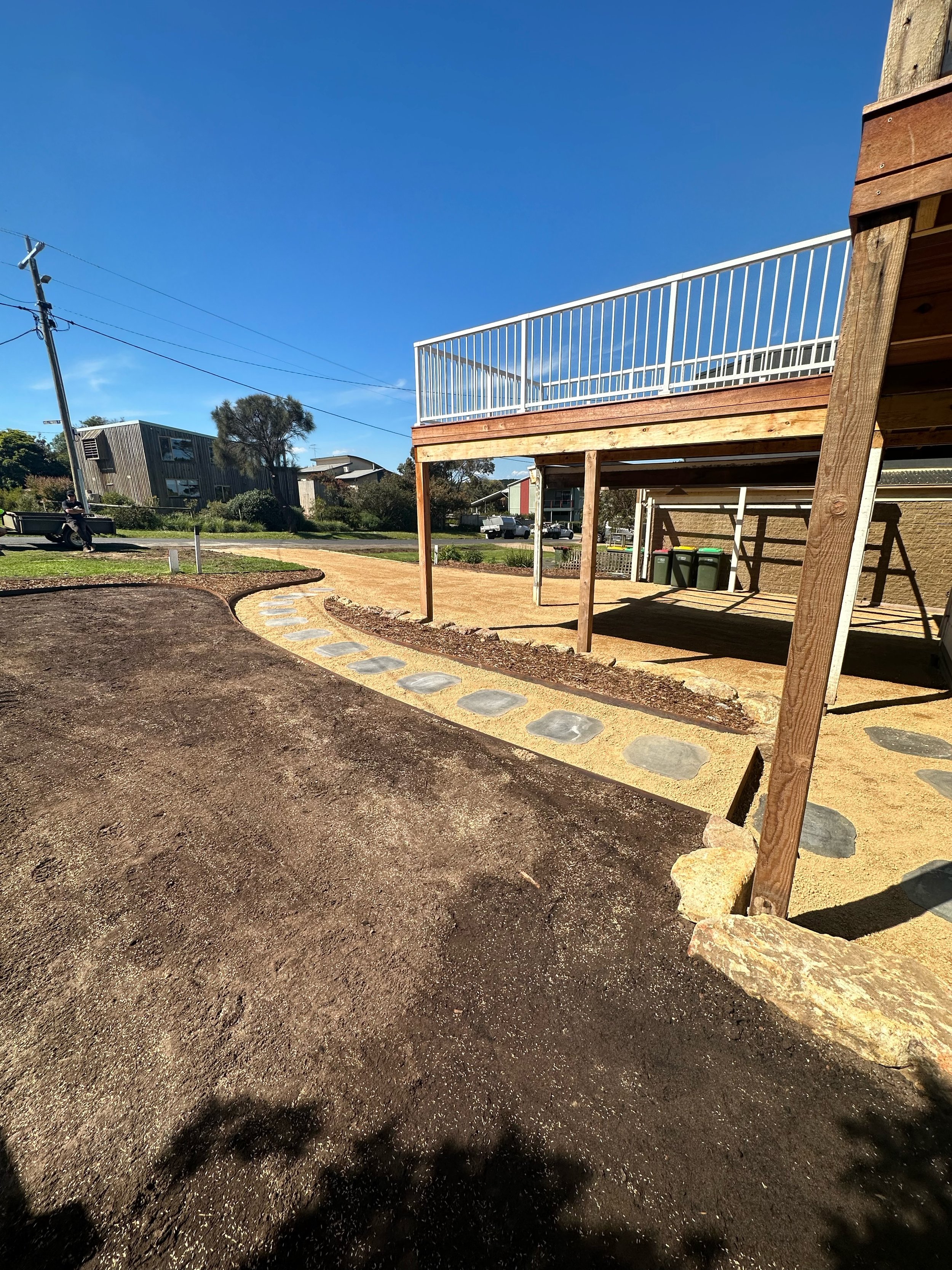 Outdoor scene with a wooden deck, gravel path, and buildings in the background under a clear blue sky.