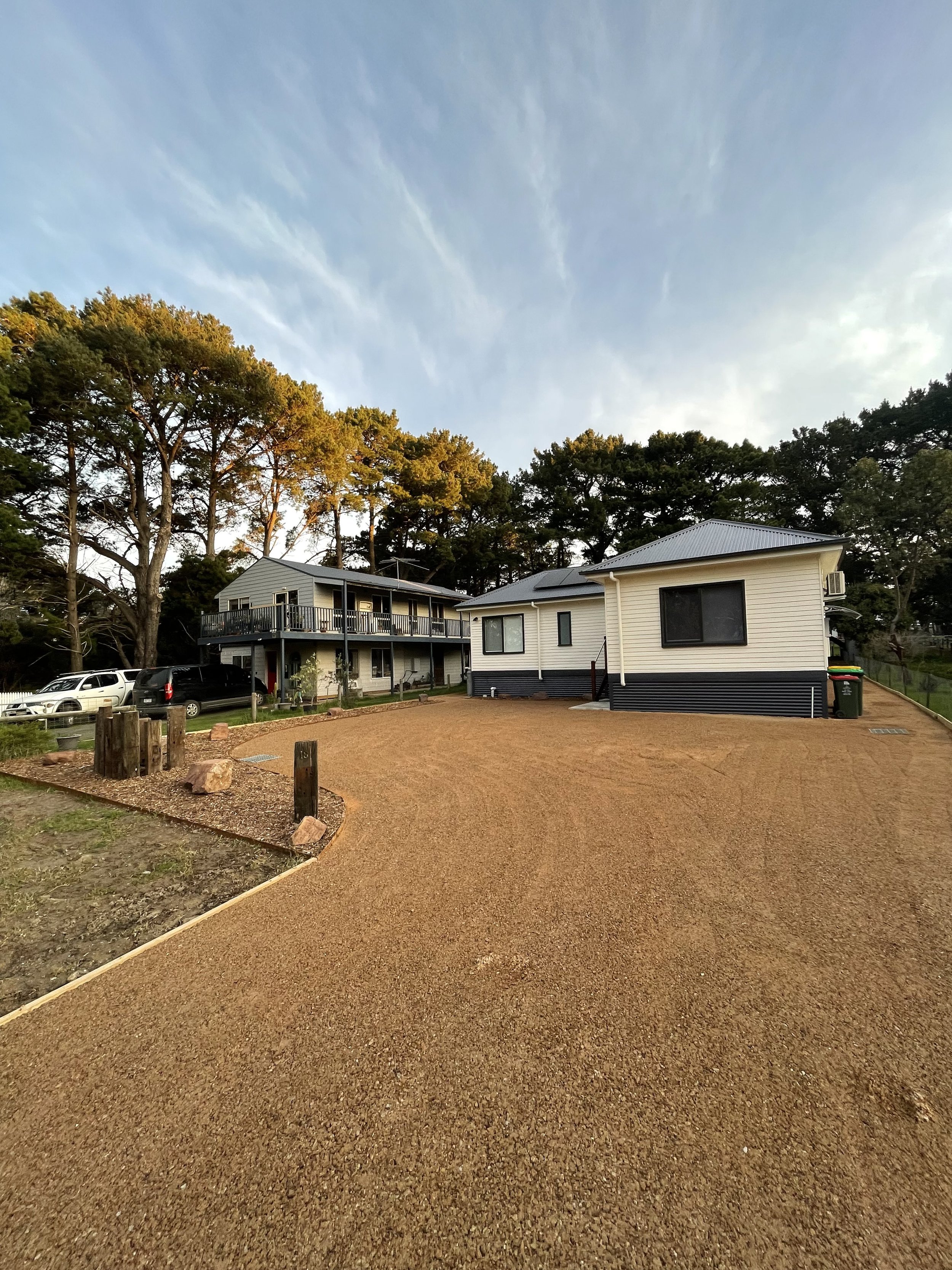 Large gravel driveway leading to two houses, surrounded by tall trees under a clear sky.