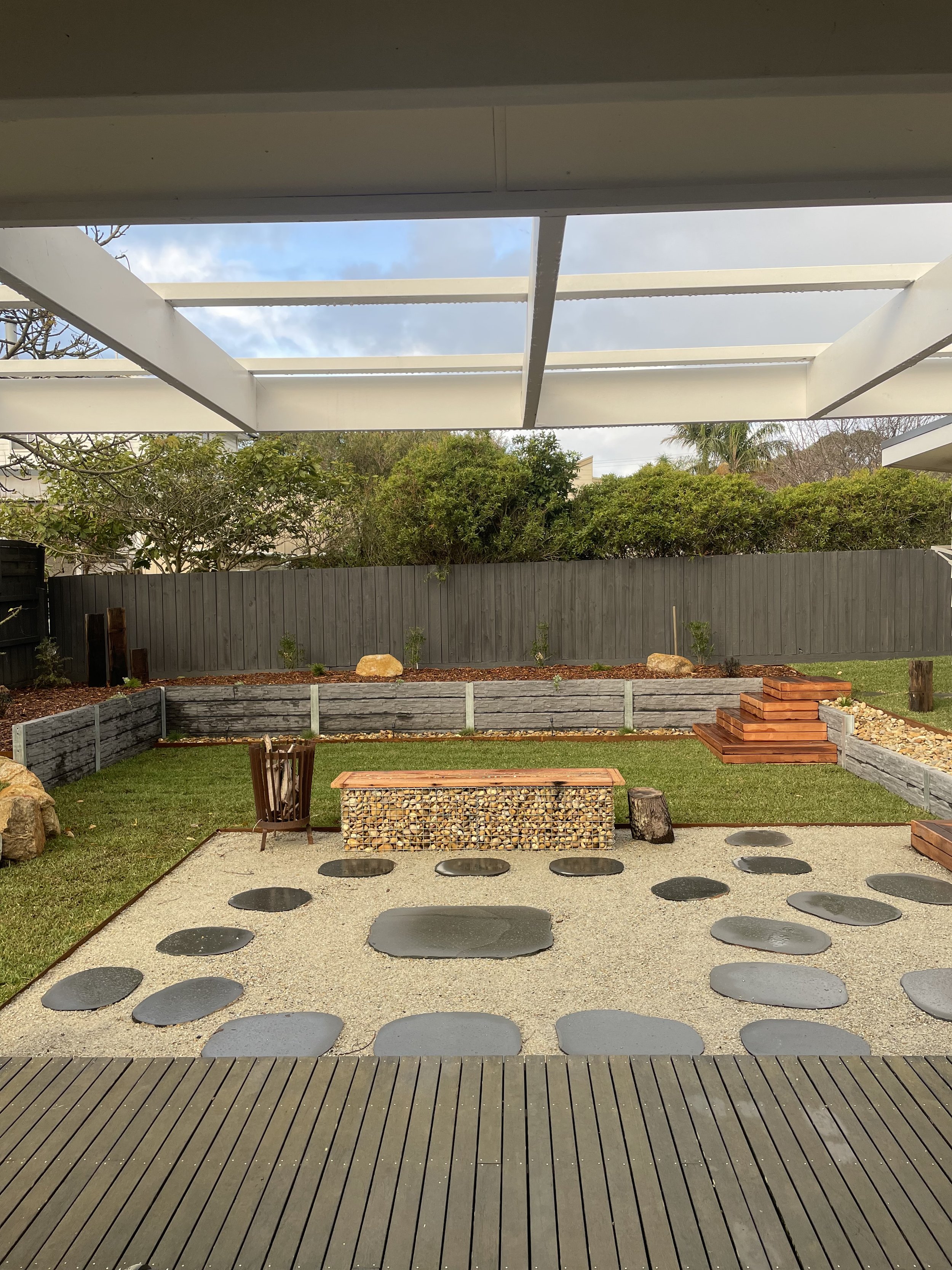 Backyard with wooden decking, stepping stones, gravel area, stone bench, grass, and wood fencing.