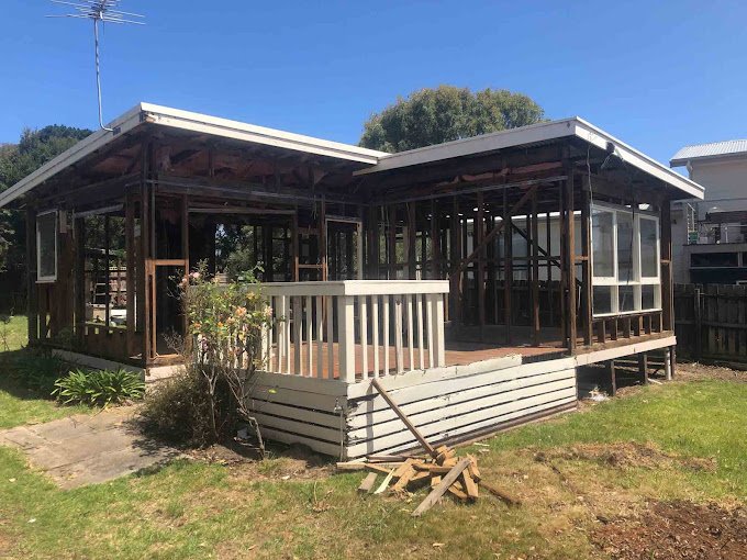 Partially constructed wooden house with exposed framework and missing walls, surrounded by grass and a few scattered planks.