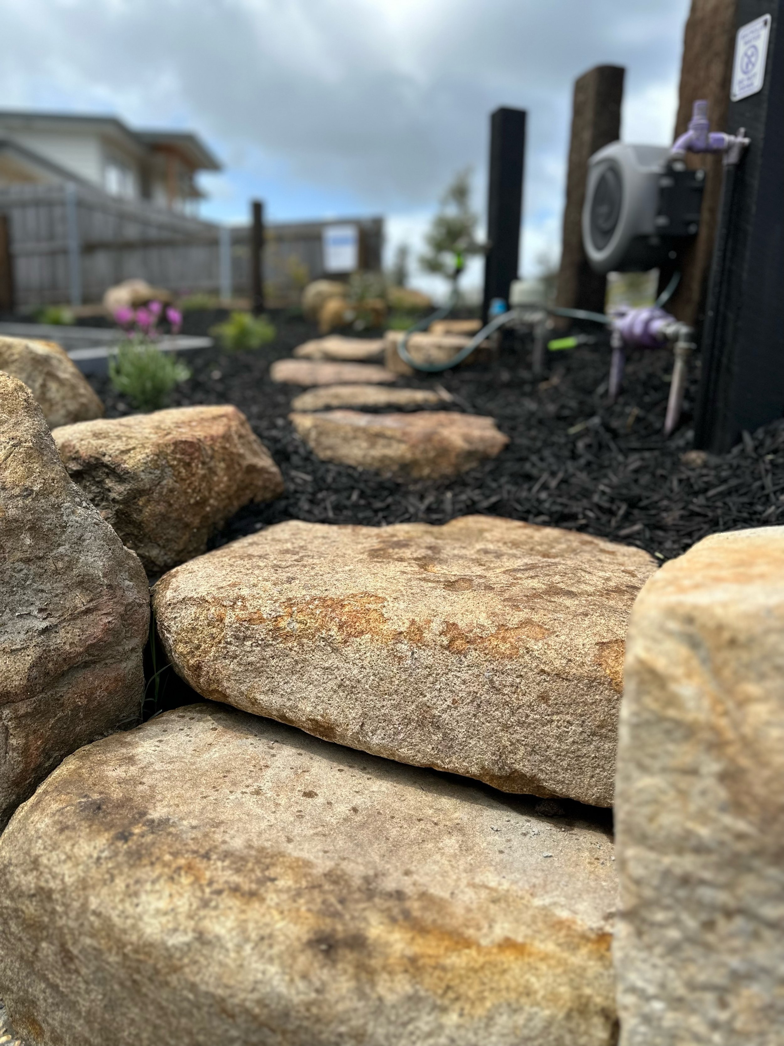 Close-up view of stone steps in a landscaped garden with mulch, plants, wooden posts, and a hose reel.