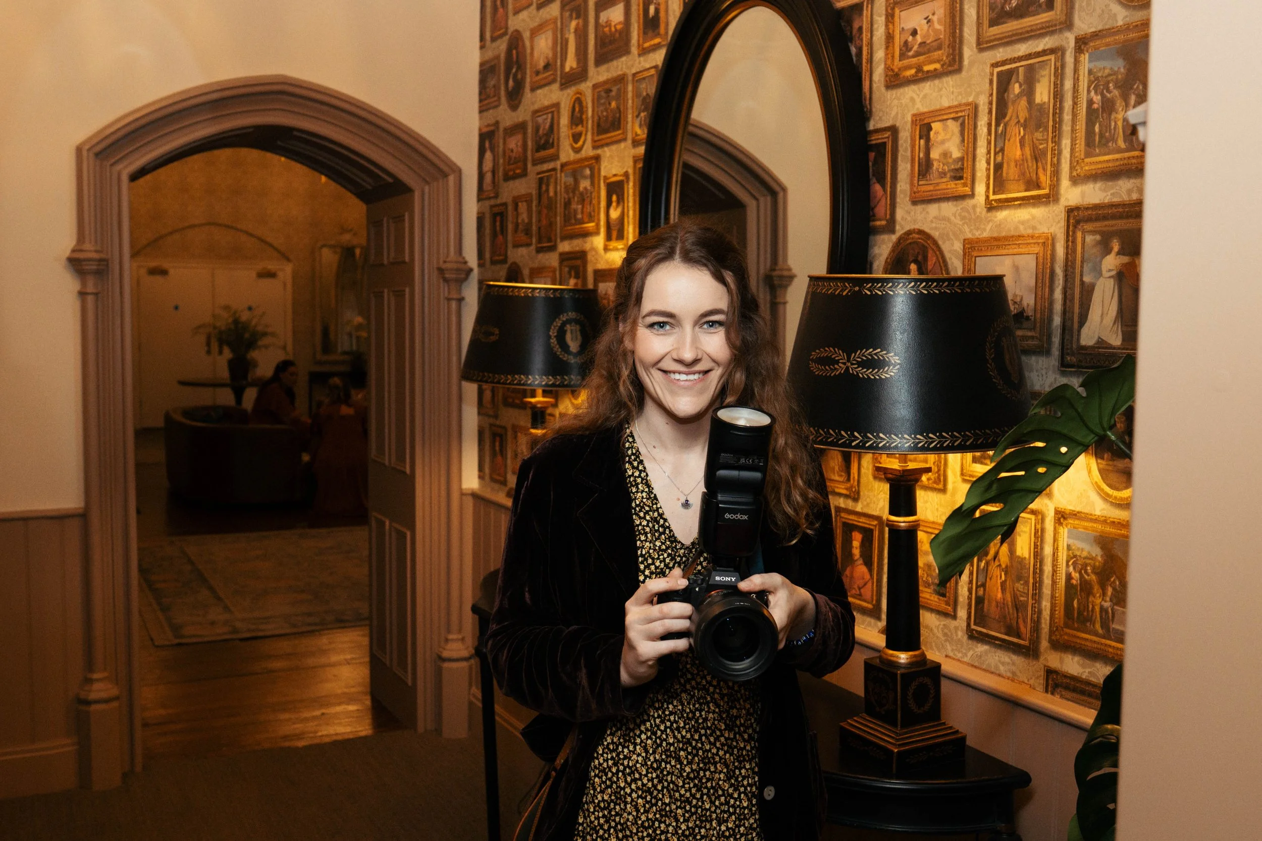 A woman with curly brown hair holding a camera, smiling, standing in front of a mirror in a room with framed artwork on the walls, lamps, and a plant.