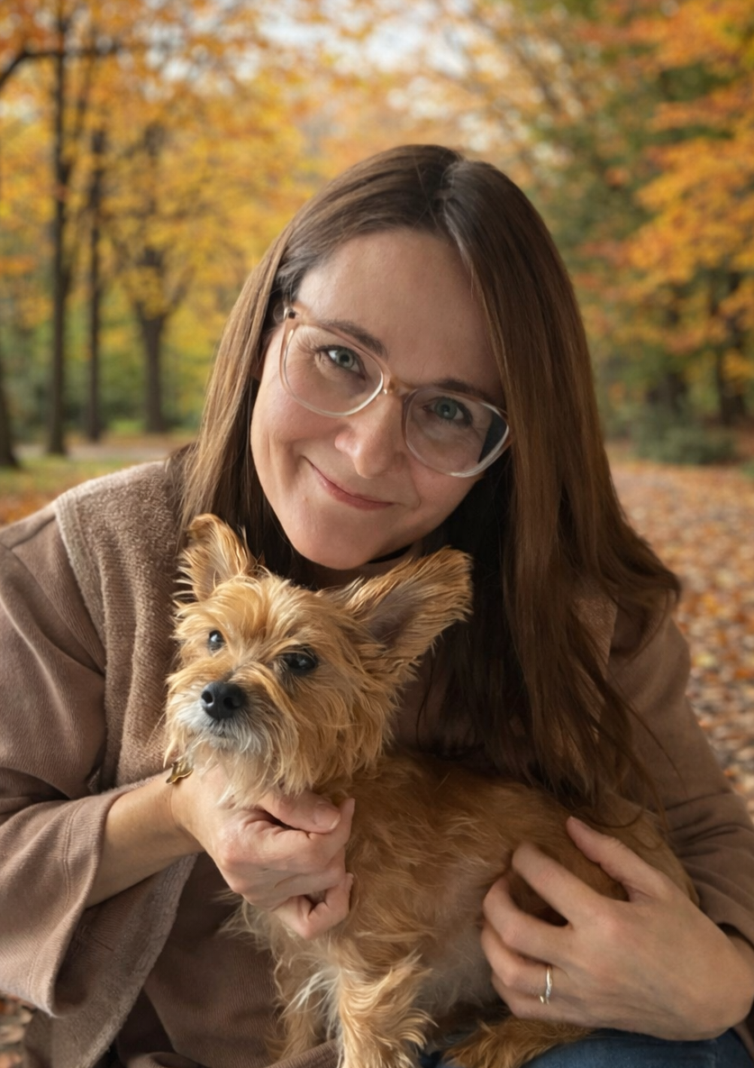 A woman with long brown hair, glasses, and a brown sweater holding a small tan-colored dog, outdoors in a park during autumn with colorful fall foliage.