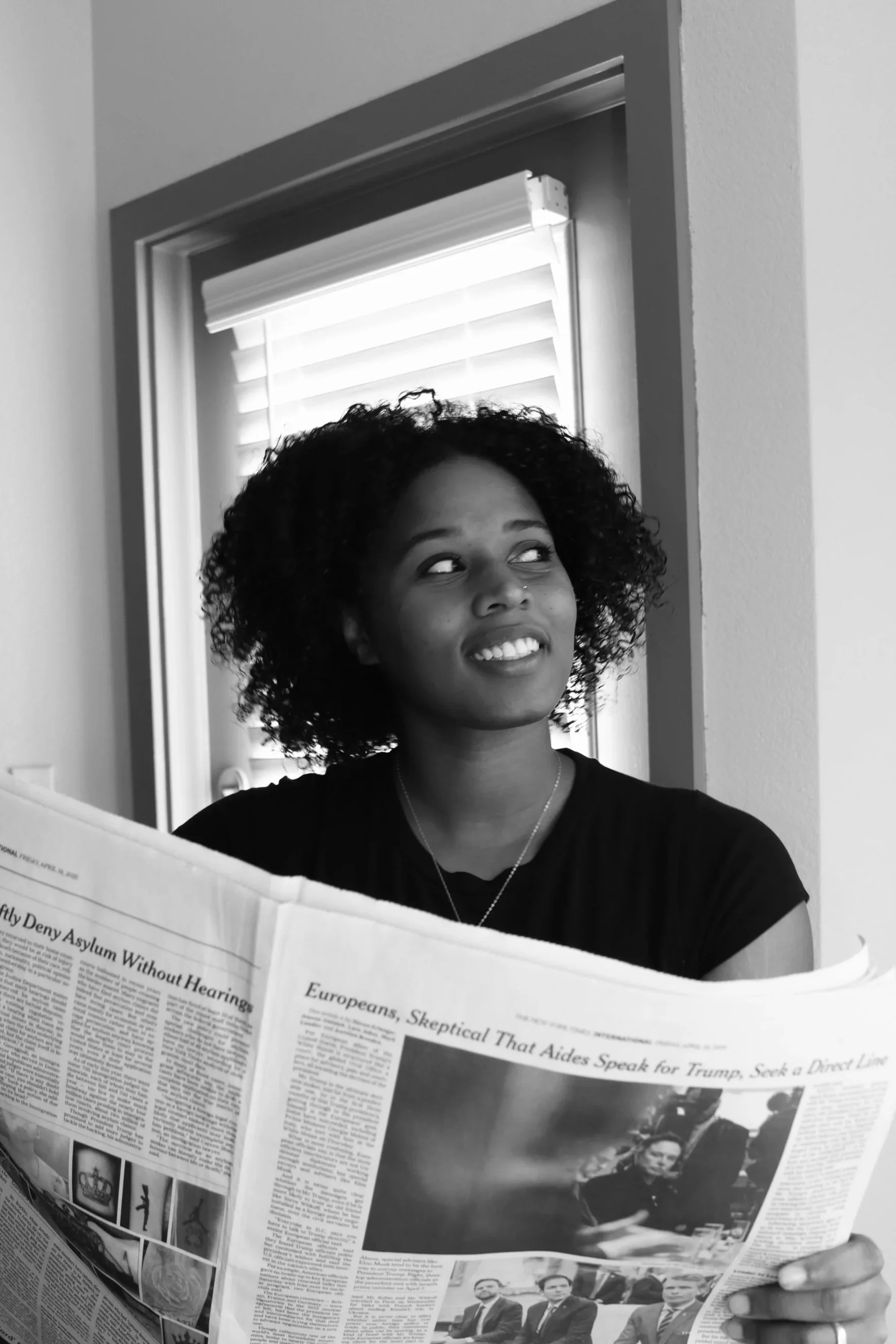 A woman with curly hair holding a newspaper, looking to the side, with a window with blinds behind her.