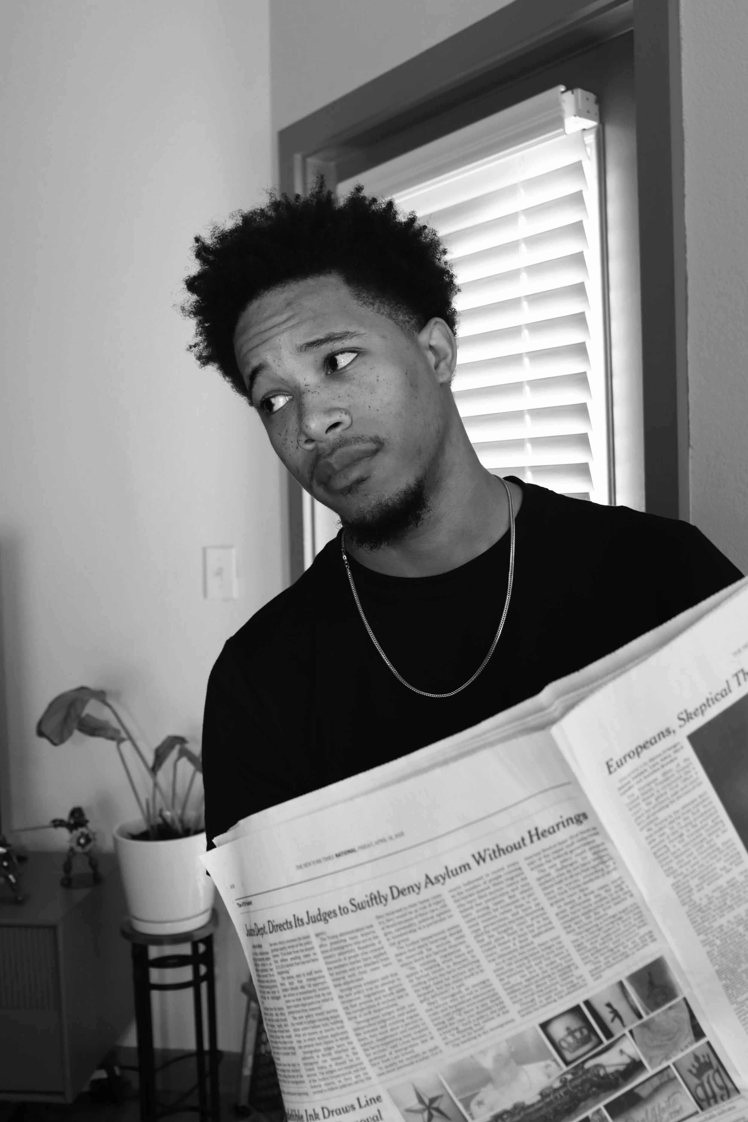 A young man with curly hair and freckles looking to the side while holding a newspaper in an indoor setting with a window with blinds and a potted plant in the background.