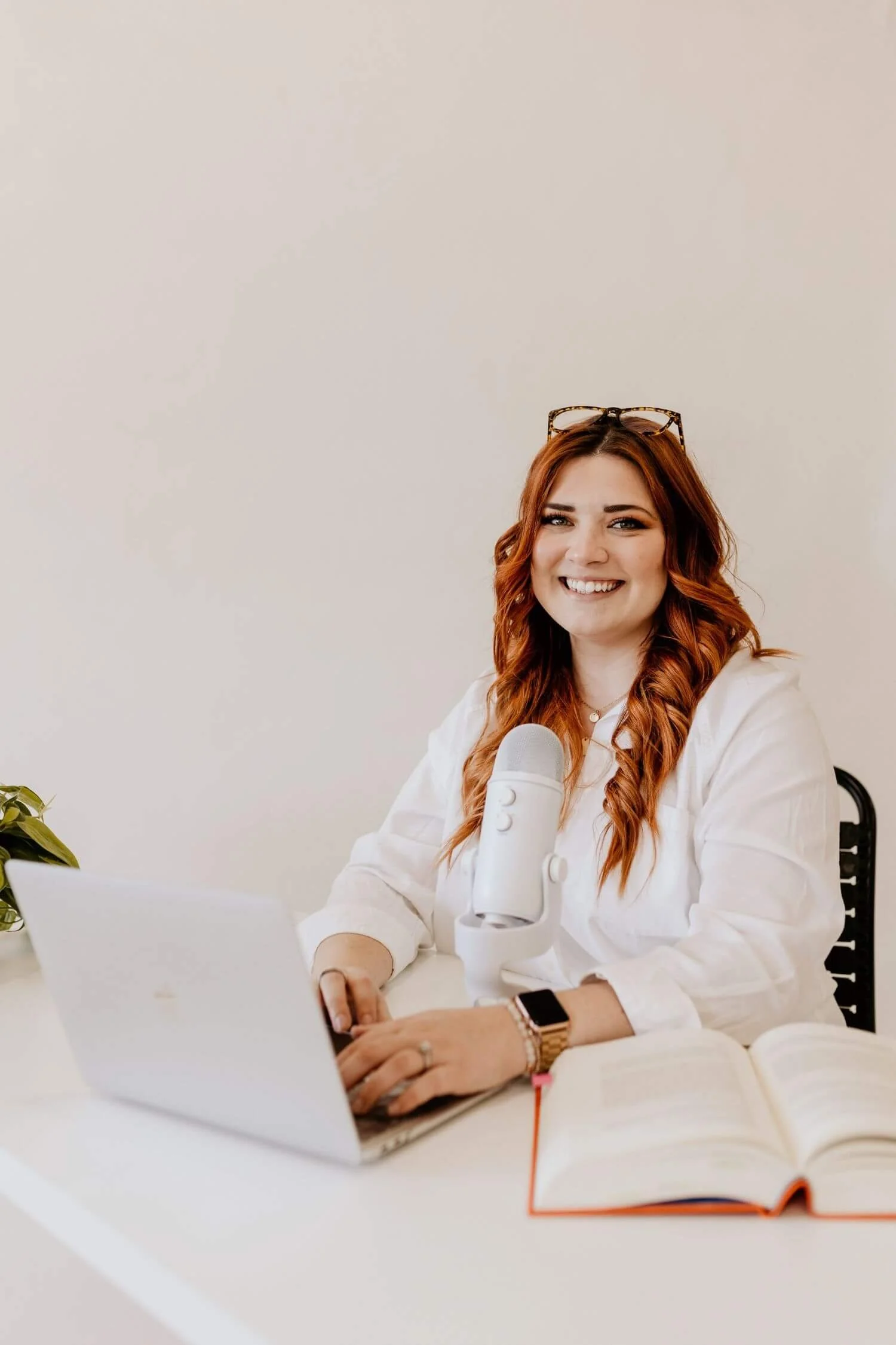 Ellen Polk wearing a white shirt, glasses on her head, and a smartwatch, sitting at a white desk with a laptop, a white microphone, an open notebook, and a potted plant, smiling directly at the camera against a plain light-colored wall.
