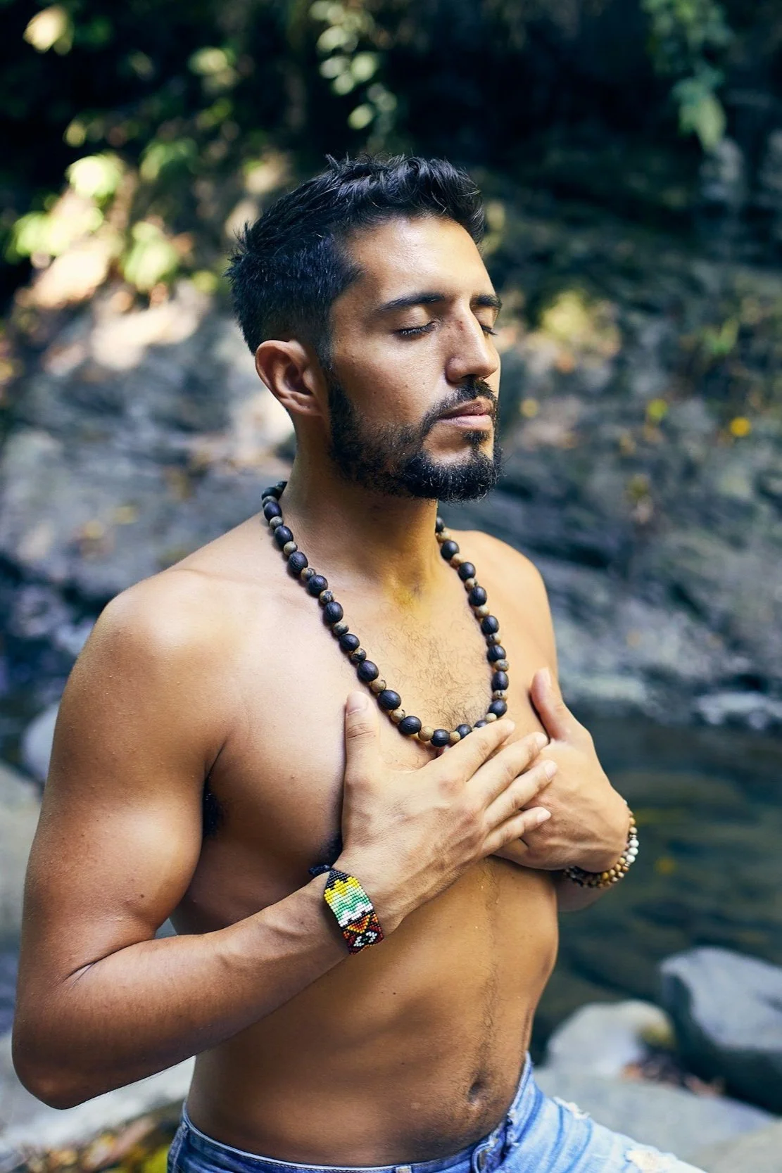 A shirtless man with closed eyes and a beard stands outdoors near rocks and trees, holding his hands over his chest, wearing a beaded necklace and bracelet.