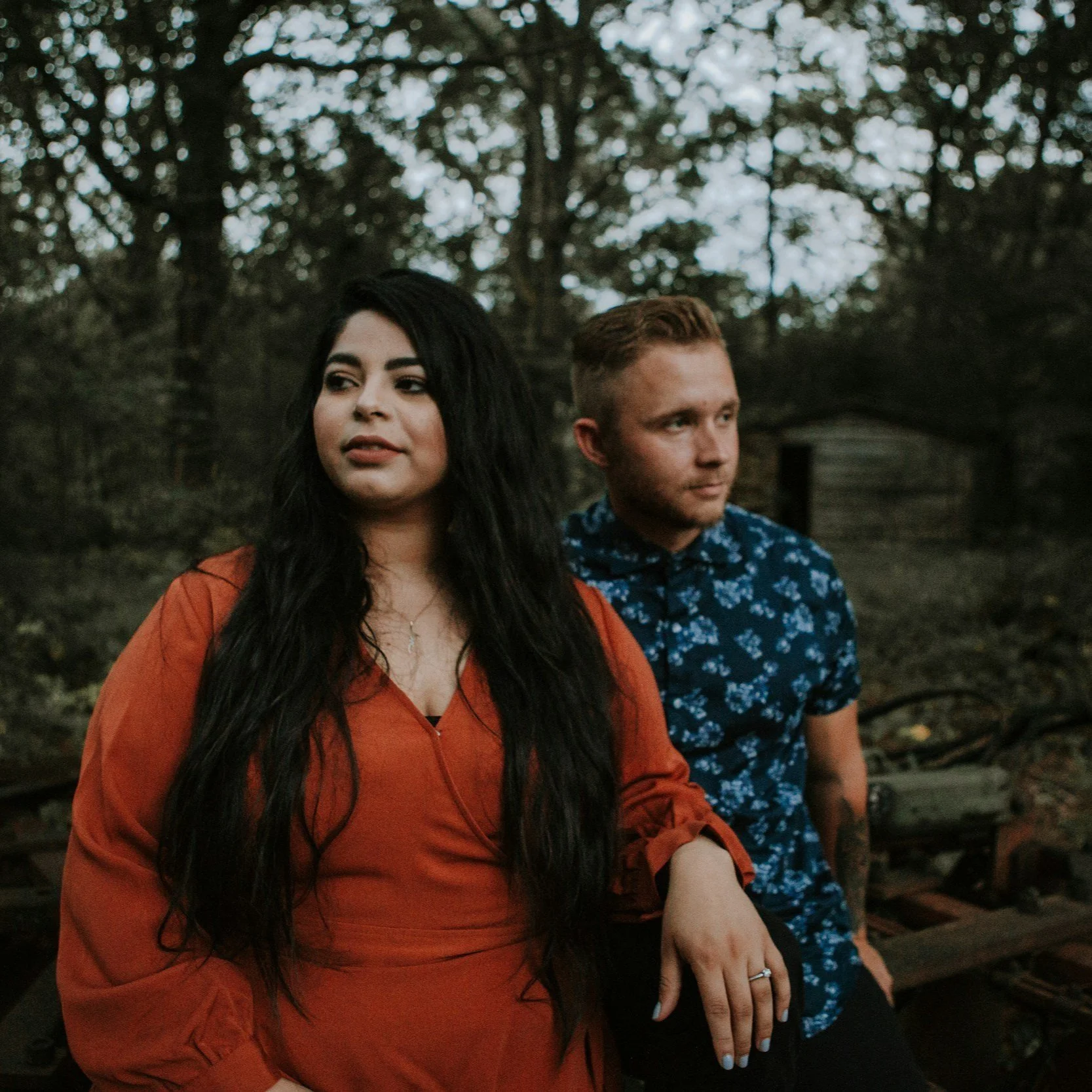 A woman and a man standing outdoors in a wooded area, with the woman in the foreground wearing an orange blouse and the man behind her wearing a blue patterned shirt.