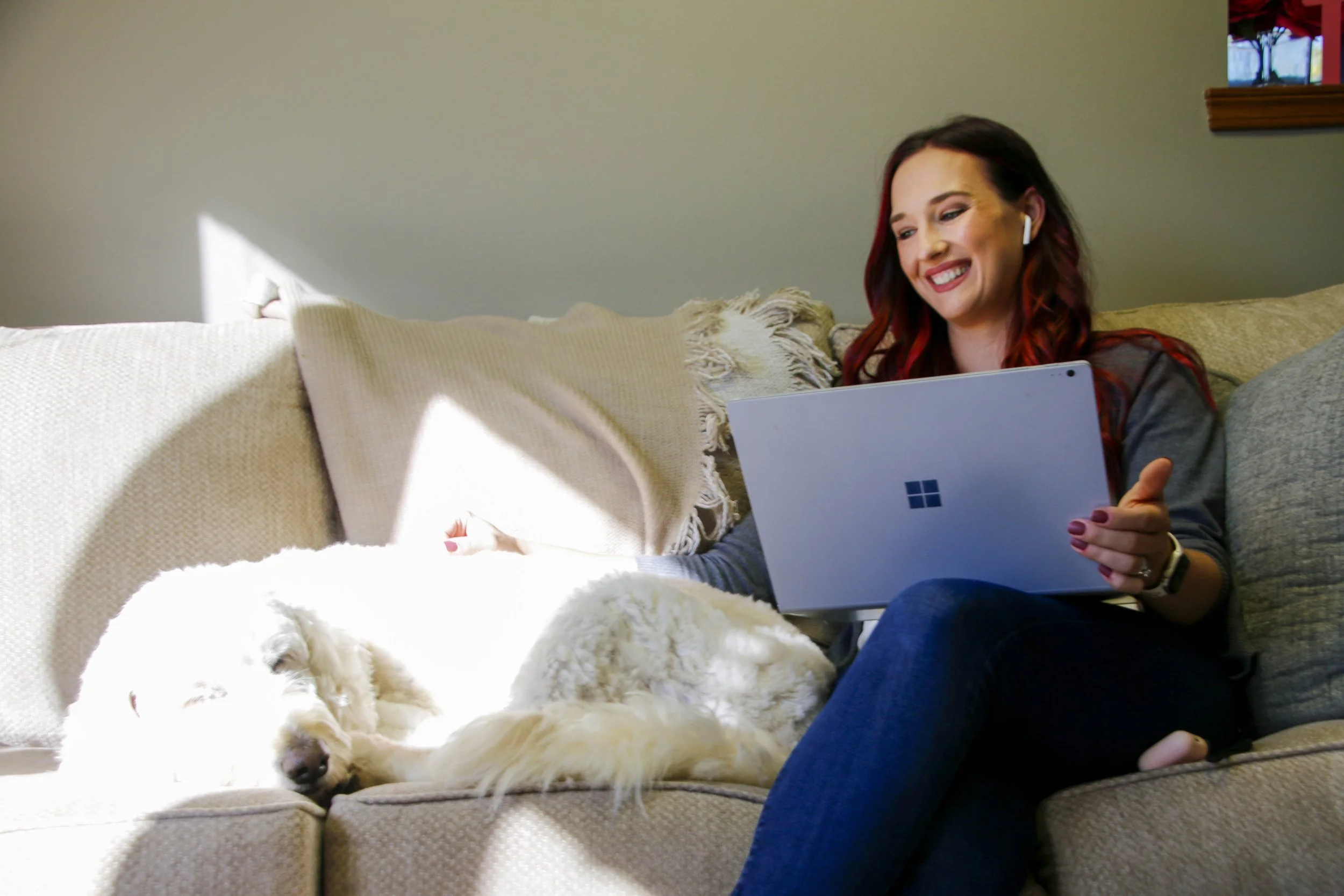A woman with red hair sitting on a beige couch, smiling while looking at a laptop with a blue Windows logo. A white dog is lying next to her, partially covered by a beige blanket.
