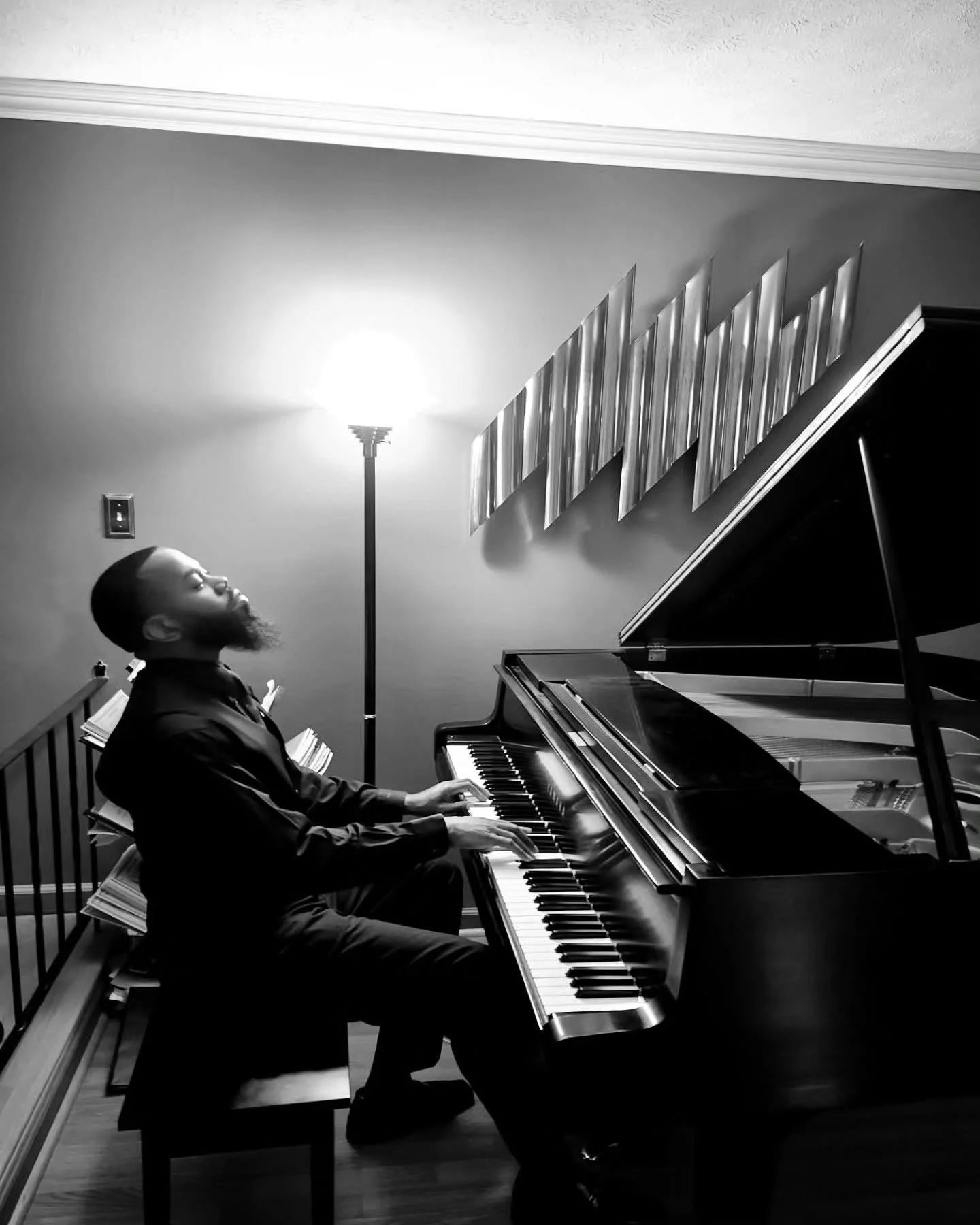 A man playing a grand piano in a room with a standing lamp and modern wall art, black and white photo.