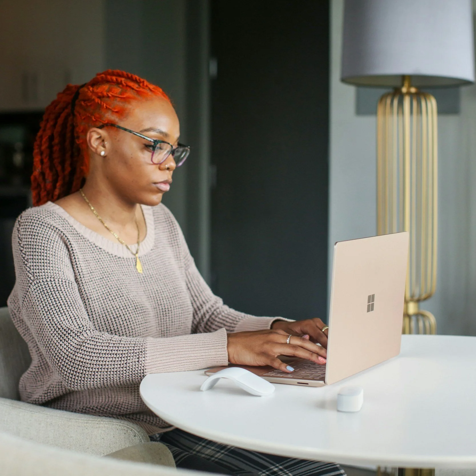 A woman with red dreadlocks, glasses, and a beige sweater working on a pink Microsoft Surface laptop at a white table. There is a white mouse and a small white device nearby, with a modern floor lamp in the background.