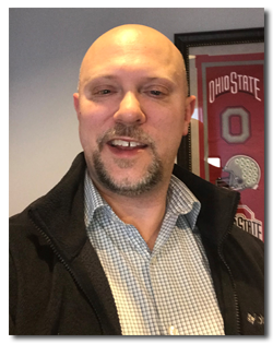 A man with a bald head, goatee, and light complexion smiling, wearing a checkered shirt and black jacket, standing indoors with an Ohio State football jersey framed on the wall behind him.