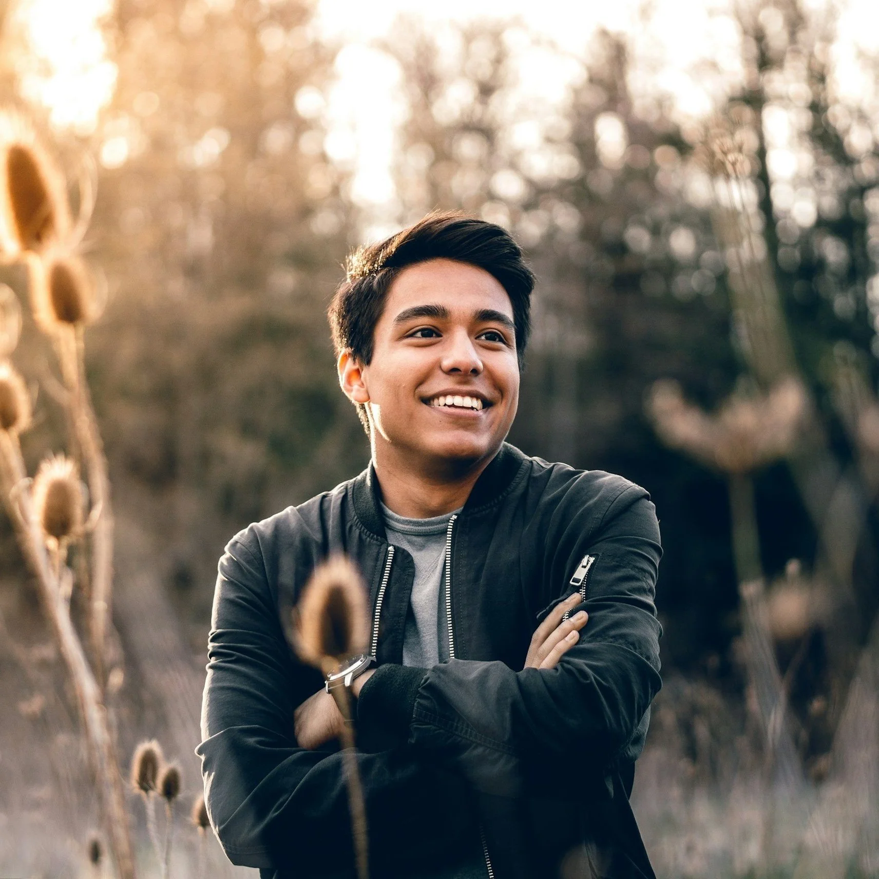 A young man smiling outdoors in a field with dry plants, with sunlight filtering through trees in the background.