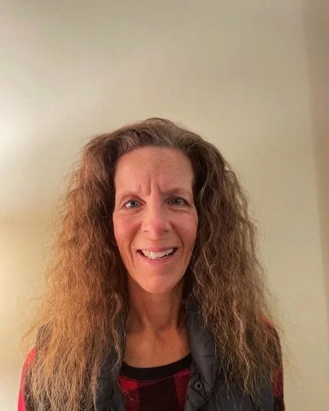 A woman with curly, frizzy hair making a puzzled or confused facial expression against a plain wall background.