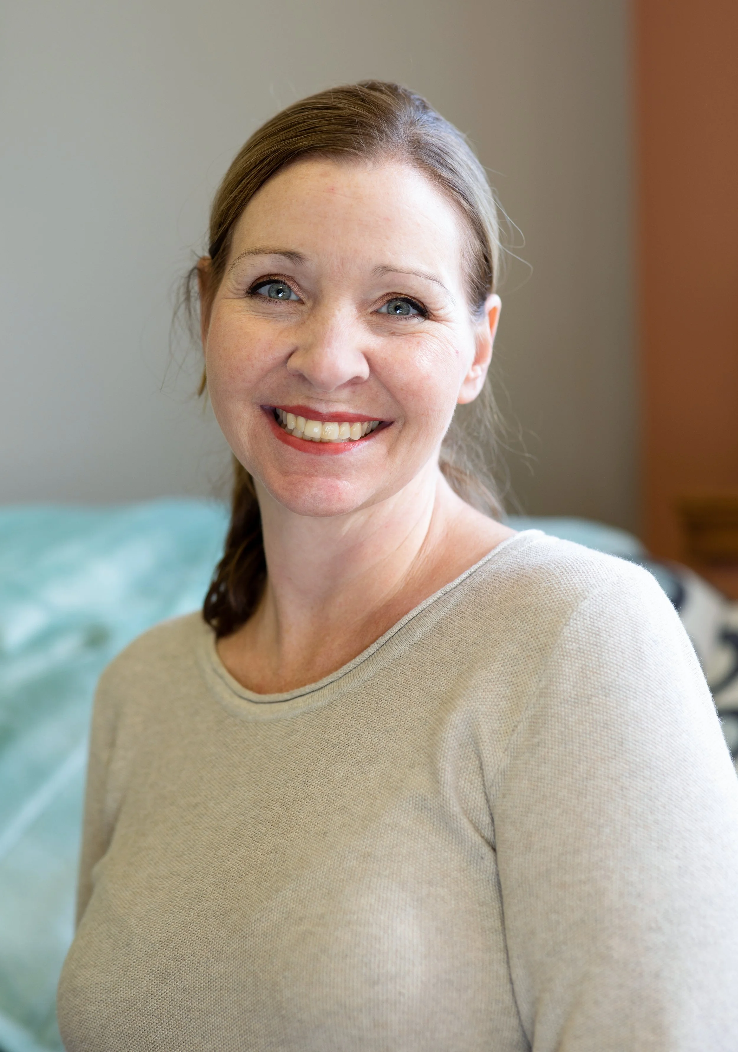 Smiling woman with light brown hair and blue eyes, wearing a beige shirt, indoors.