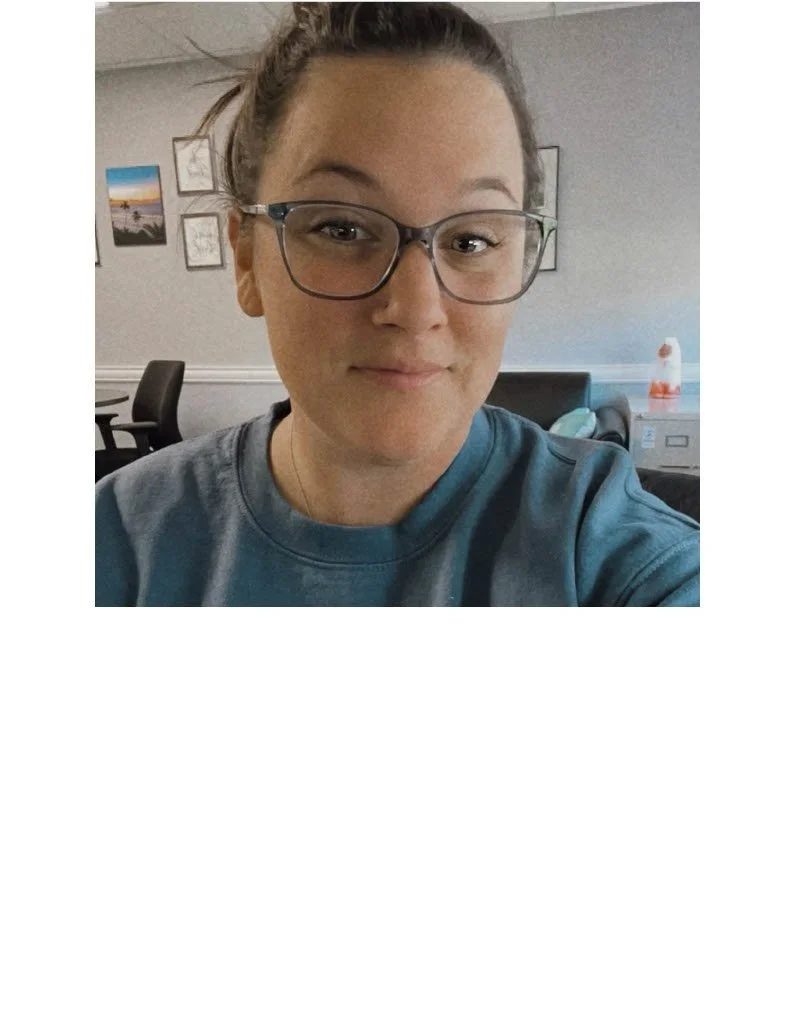 A woman with glasses and tied-back hair takes a selfie in an office, with framed pictures on the wall and a desk with a lamp in the background.