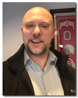Man taking a selfie in front of Ohio State University memorabilia, bald with a beard, wearing a checkered shirt and dark jacket.