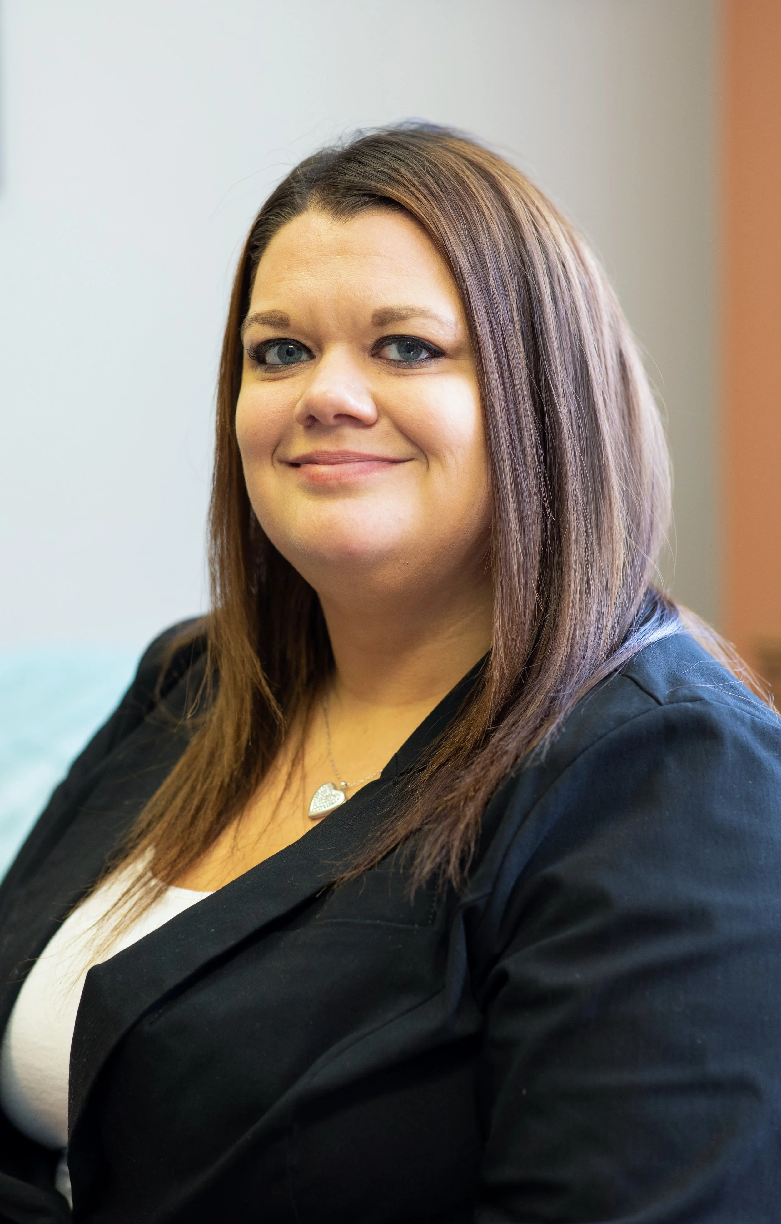 Portrait of a smiling woman with shoulder-length brown hair and blue eyes, wearing a black blazer and a heart-shaped necklace