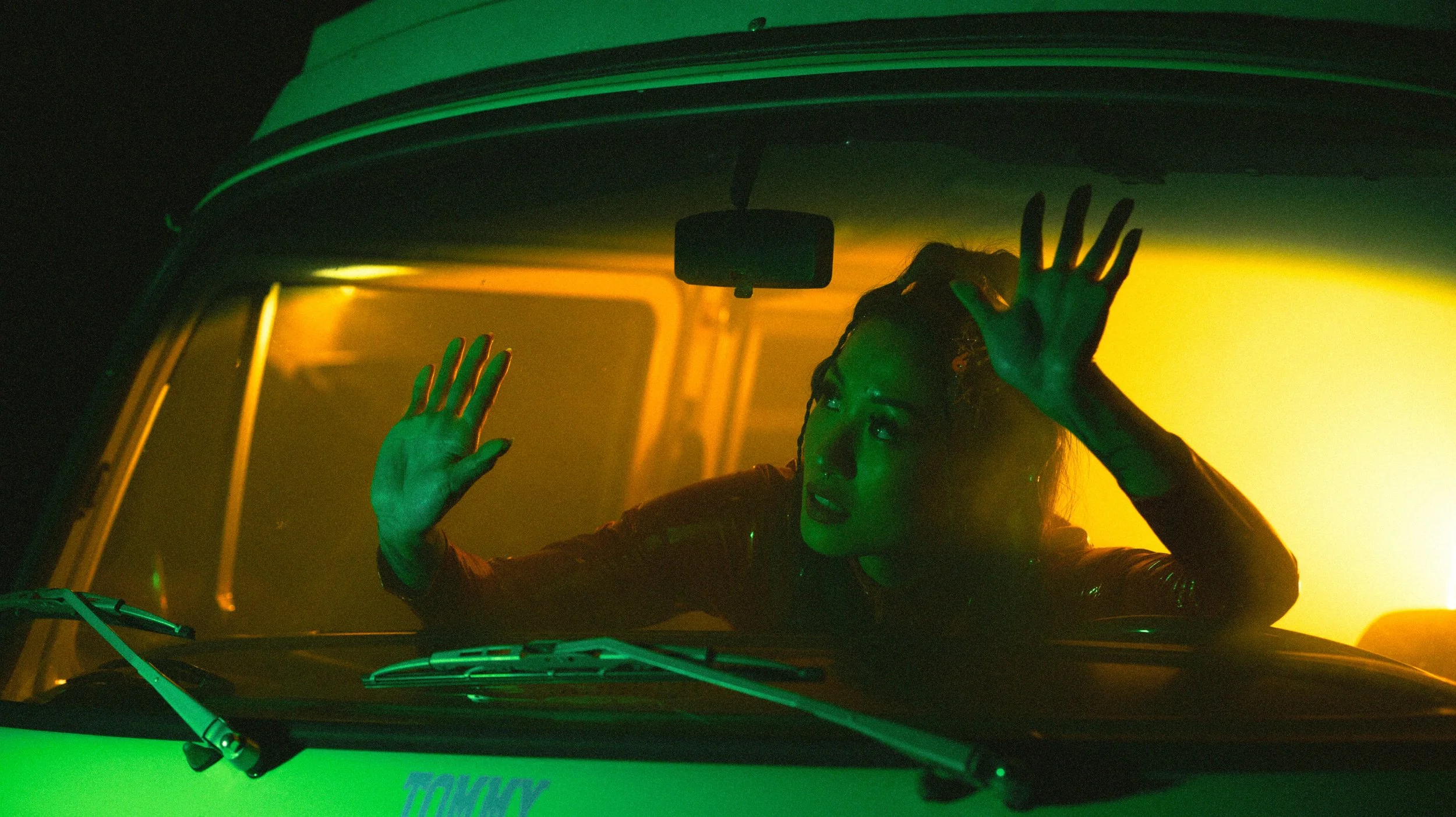 A woman with curly hair sitting in the driver's seat of a vehicle, seen through the windshield, with her hands pressed against the glass and looking to the side, illuminated by green and yellow lighting.