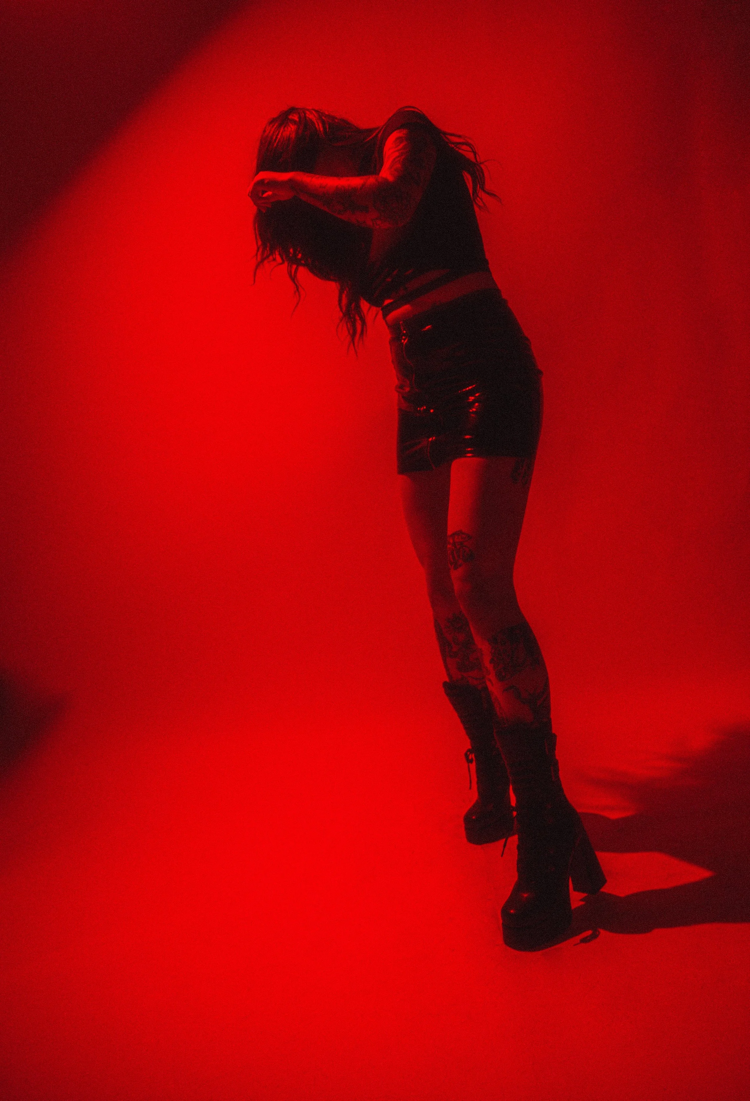 A person with tattoos is standing against a red background, covering their face with their hands, wearing a black top, a black shiny skirt, and black platform boots.