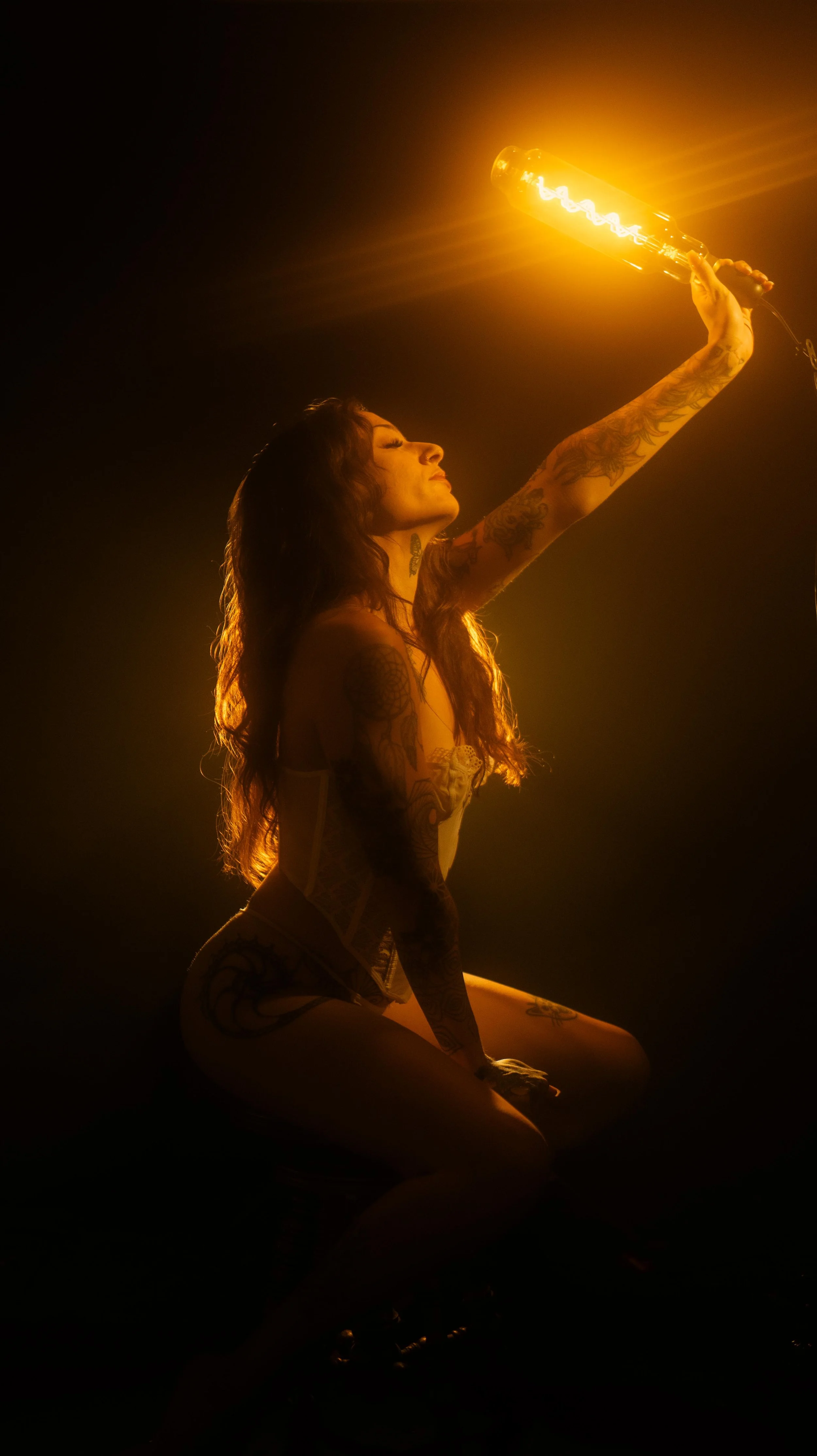 Tattooed woman in lingerie holding glowing light bulb, artistic lighting