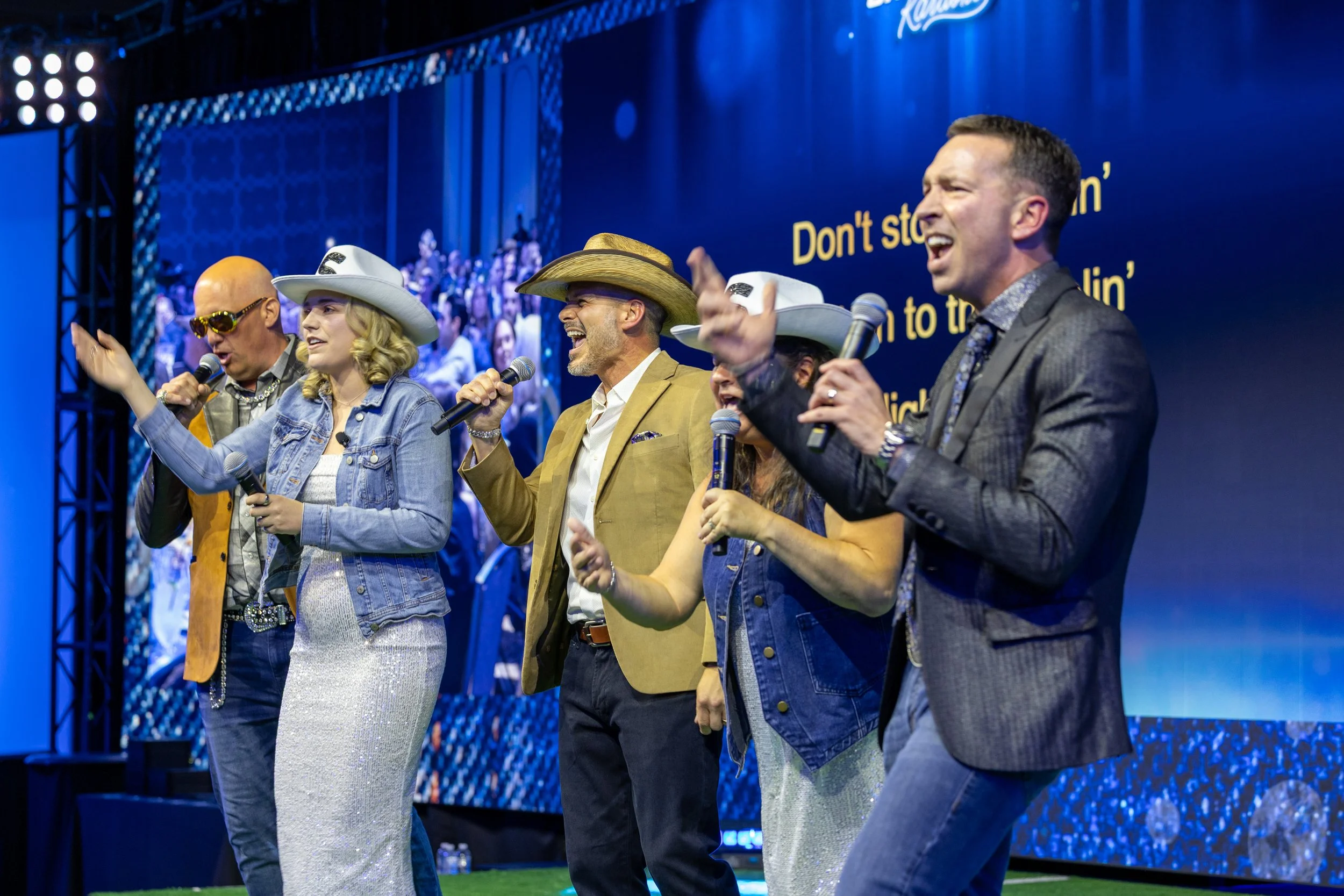 A high-energy group of corporate employees singing karaoke on stage with microphones during a Western-themed evening celebration at a Dallas-Fort Worth company event.