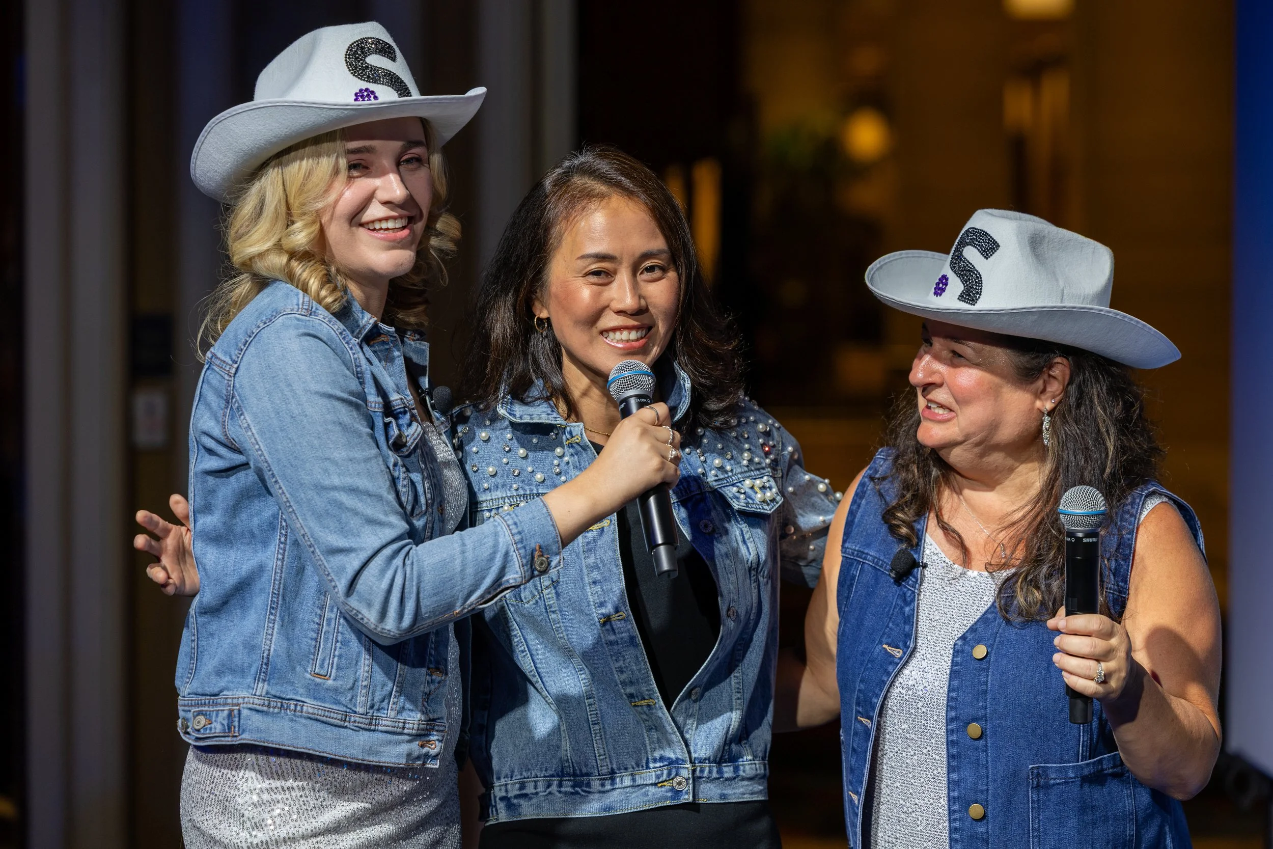 Three female corporate presenters smiling and holding microphones on stage during a Texas-themed awards gala and team-building event in the Dallas-Fort Worth area.