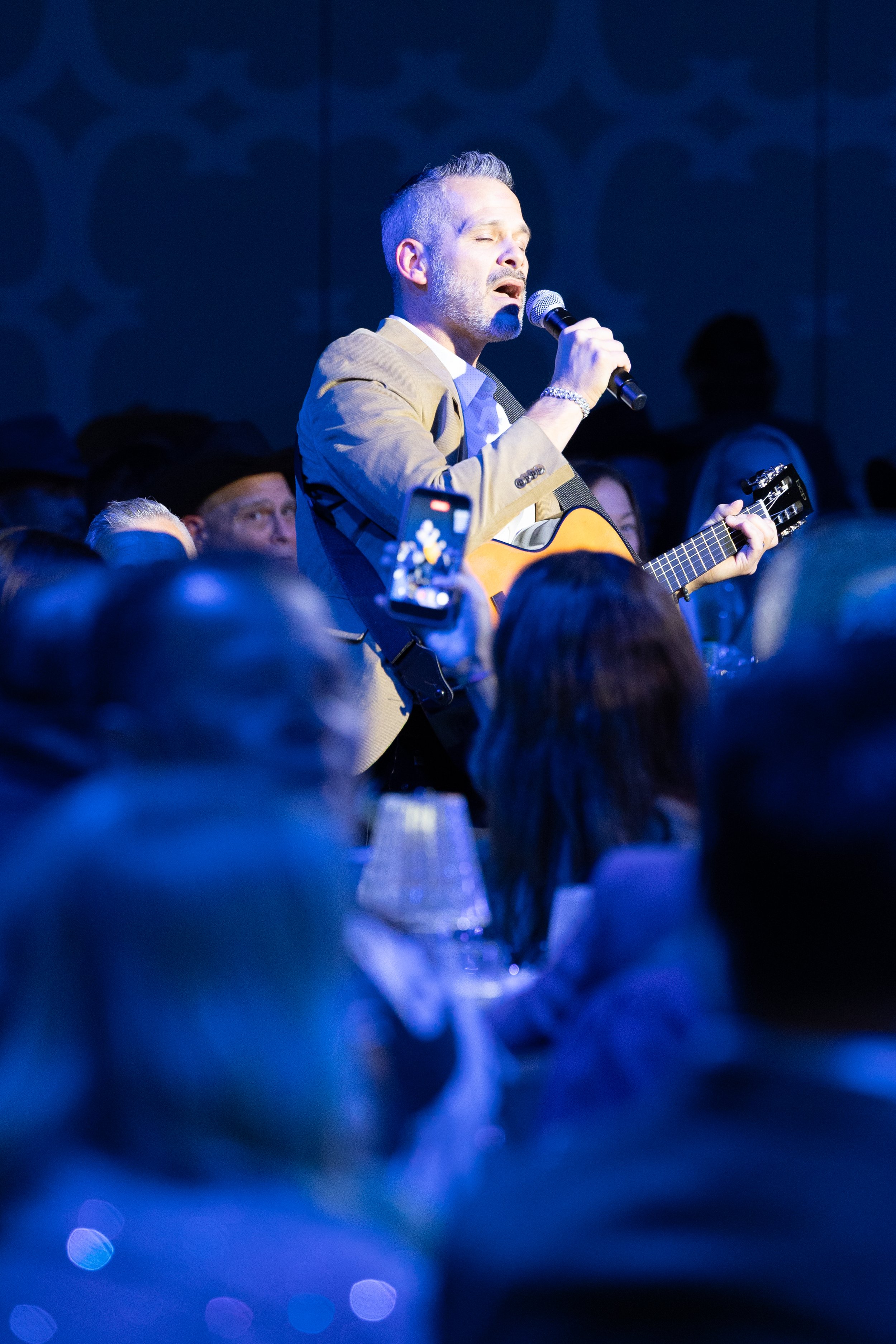 A live musical performer singing and playing an acoustic guitar under dramatic blue stage lighting during a corporate gala banquet dinner in Dallas-Fort Worth.