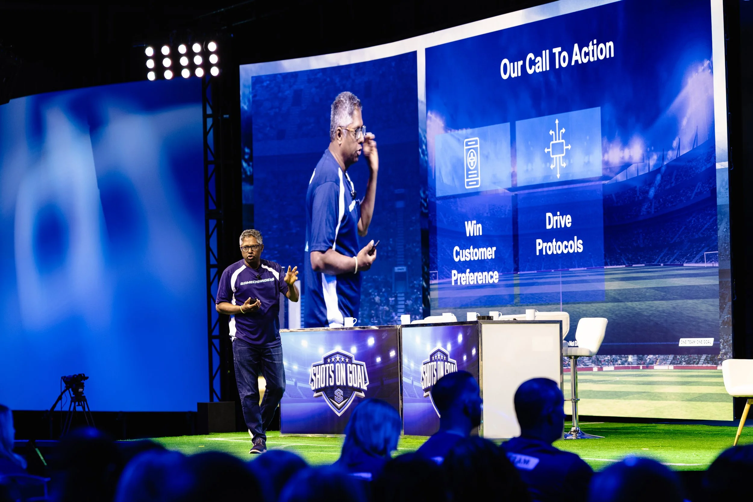 A speaker presenting on a massive convention stage with large LED screens showing presentation slides and live video feeds at a corporate seminar in the Dallas-Fort Worth area.