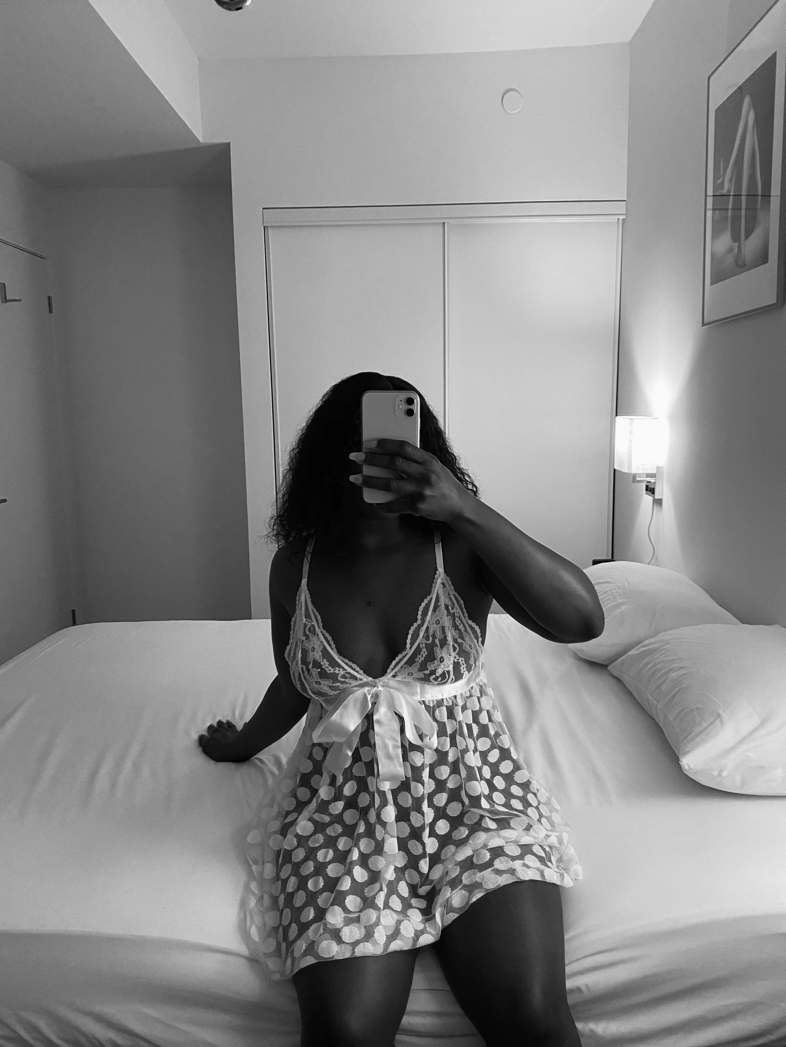 A woman taking a mirror selfie in a bedroom, sitting on a bed with white sheets, wearing a polka dot dress with lace details.
