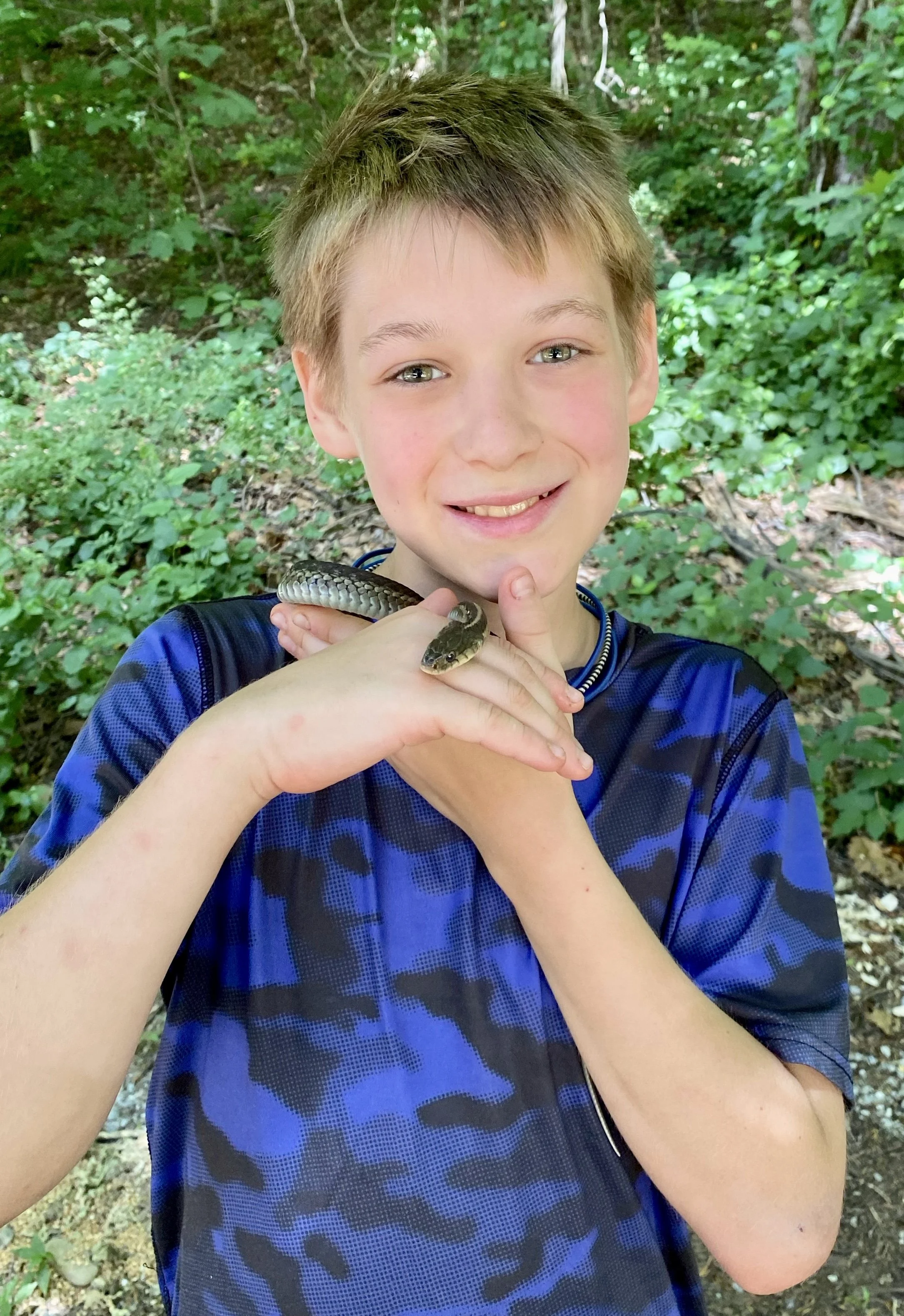 A smiling young boy wearing a blue camouflage shirt holding a small snake on his hand in a green outdoor forest setting.
