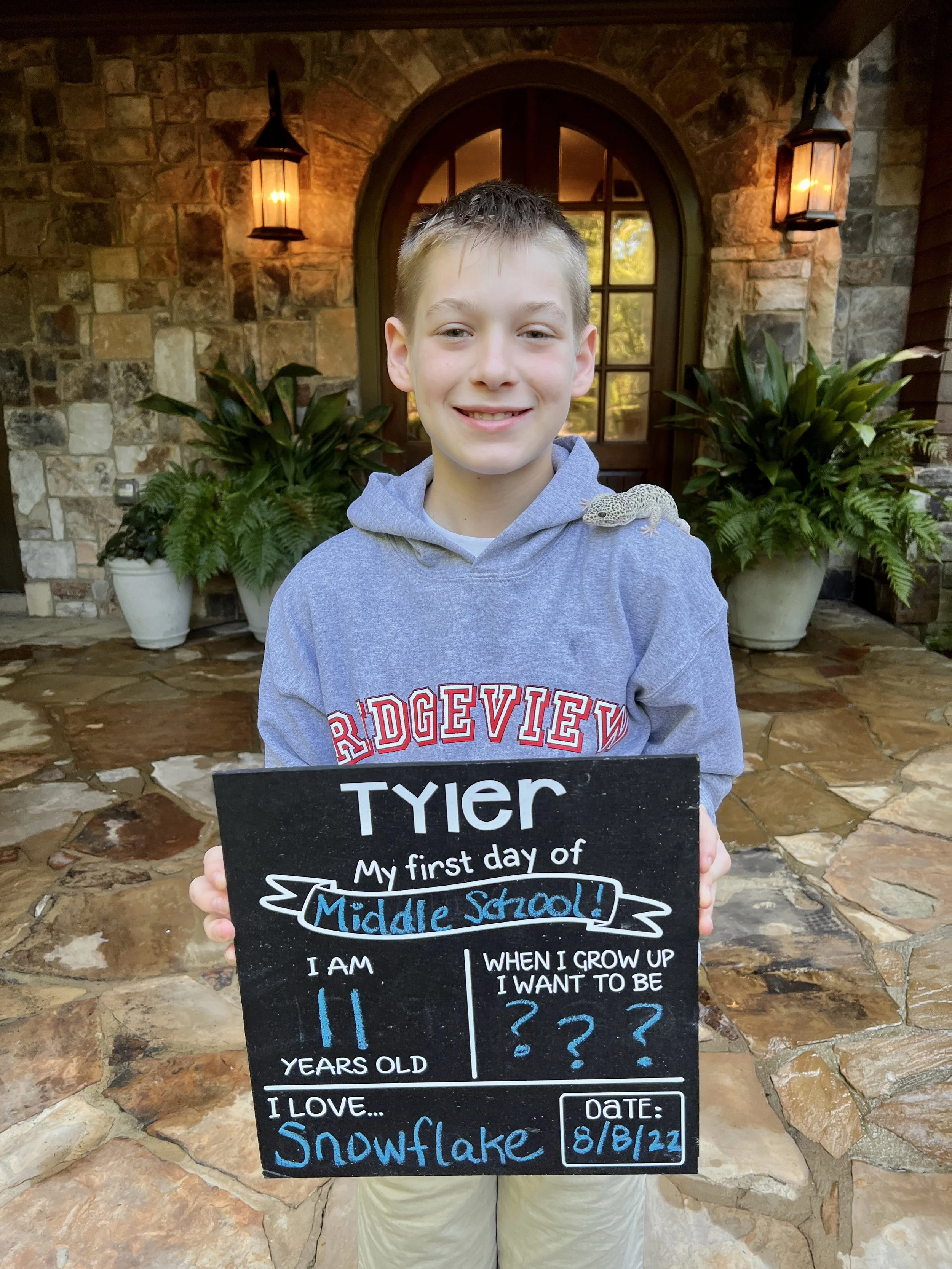 A young boy named Tyler holding a sign about his first day of middle school, standing outside in front of a stone wall with potted plants and lanterns.