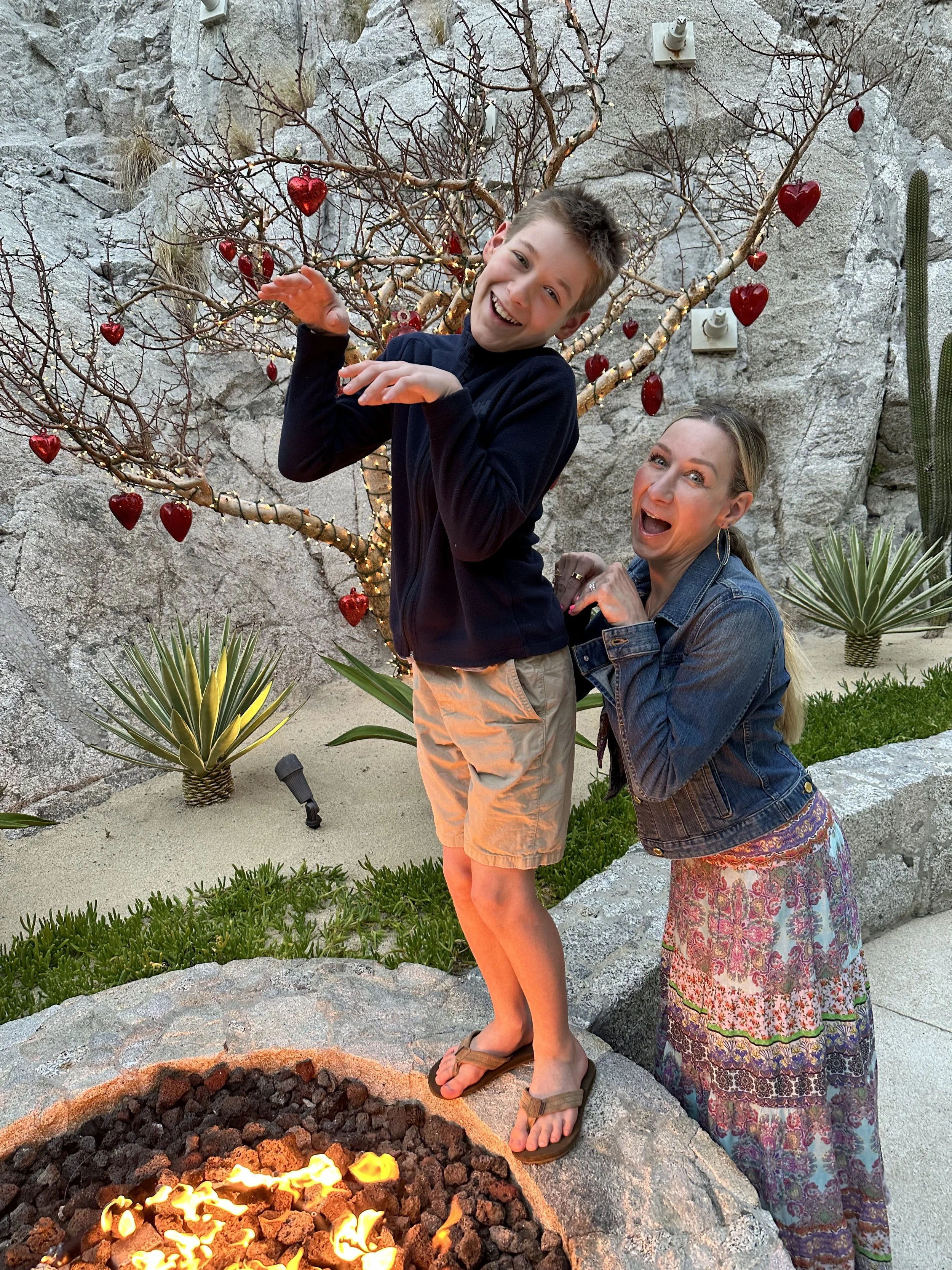 A boy standing on a rock with a woman behind him, posing playfully next to a fire pit with glowing flames. They are outdoors in front of a decorative tree with red heart-shaped ornaments and string lights, surrounded by desert plants and rocks.