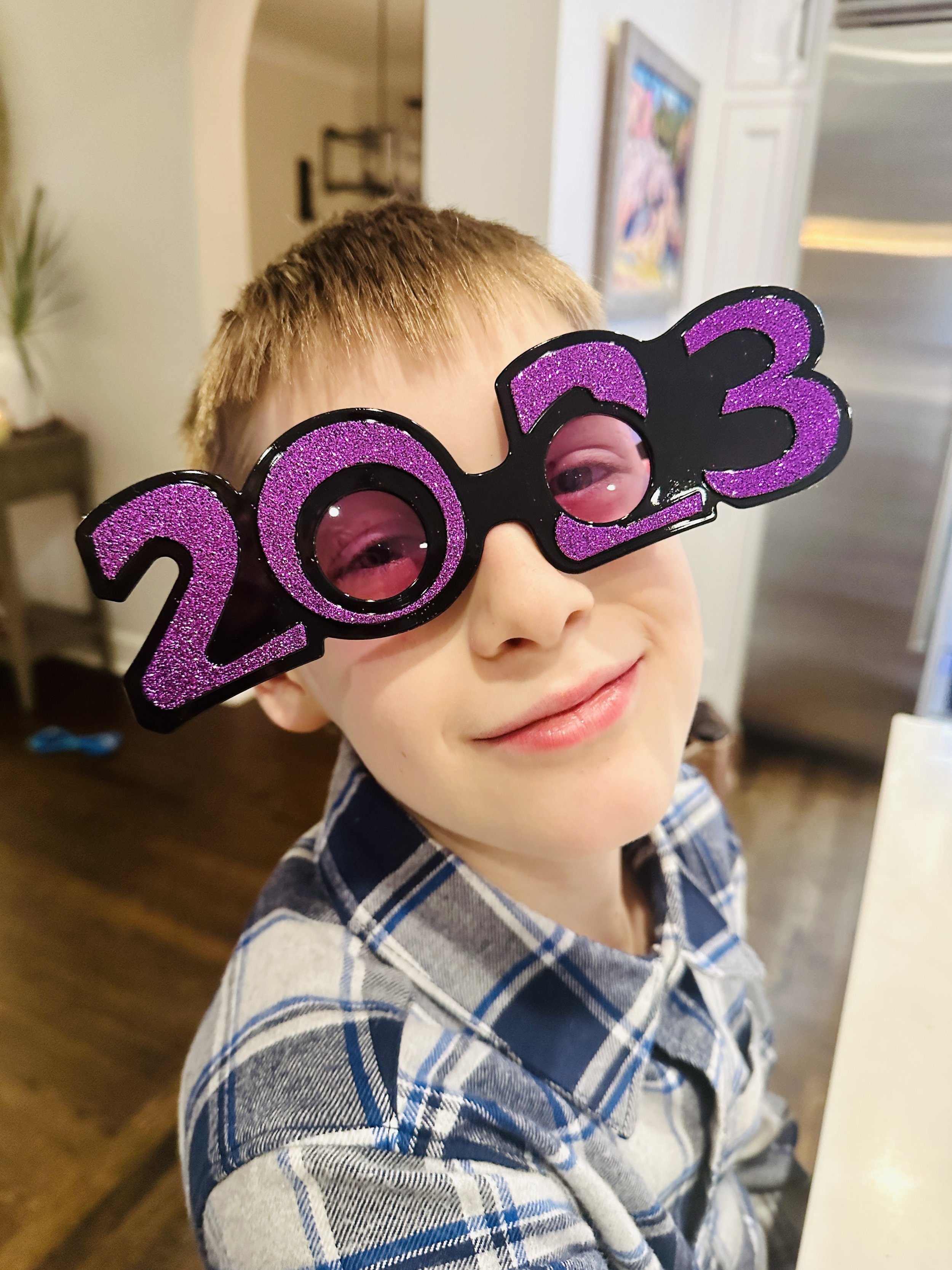 A smiling young boy wearing glasses that spell out 2023 in purple glitter, celebrating New Year's.