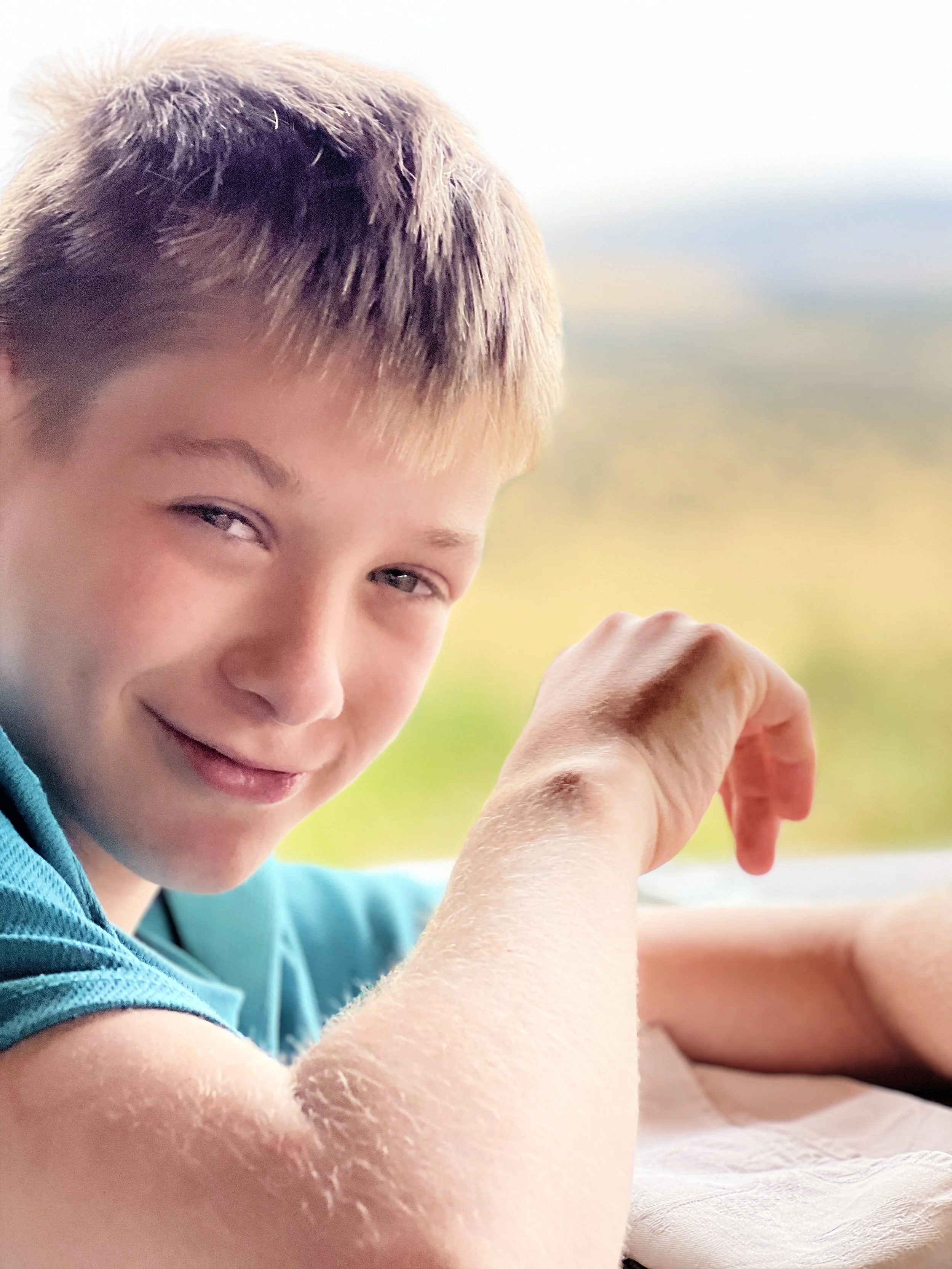 Close-up of a smiling boy with short, light brown hair, wearing a blue shirt, sitting outdoors with a green landscape and blue sky blurry in the background.