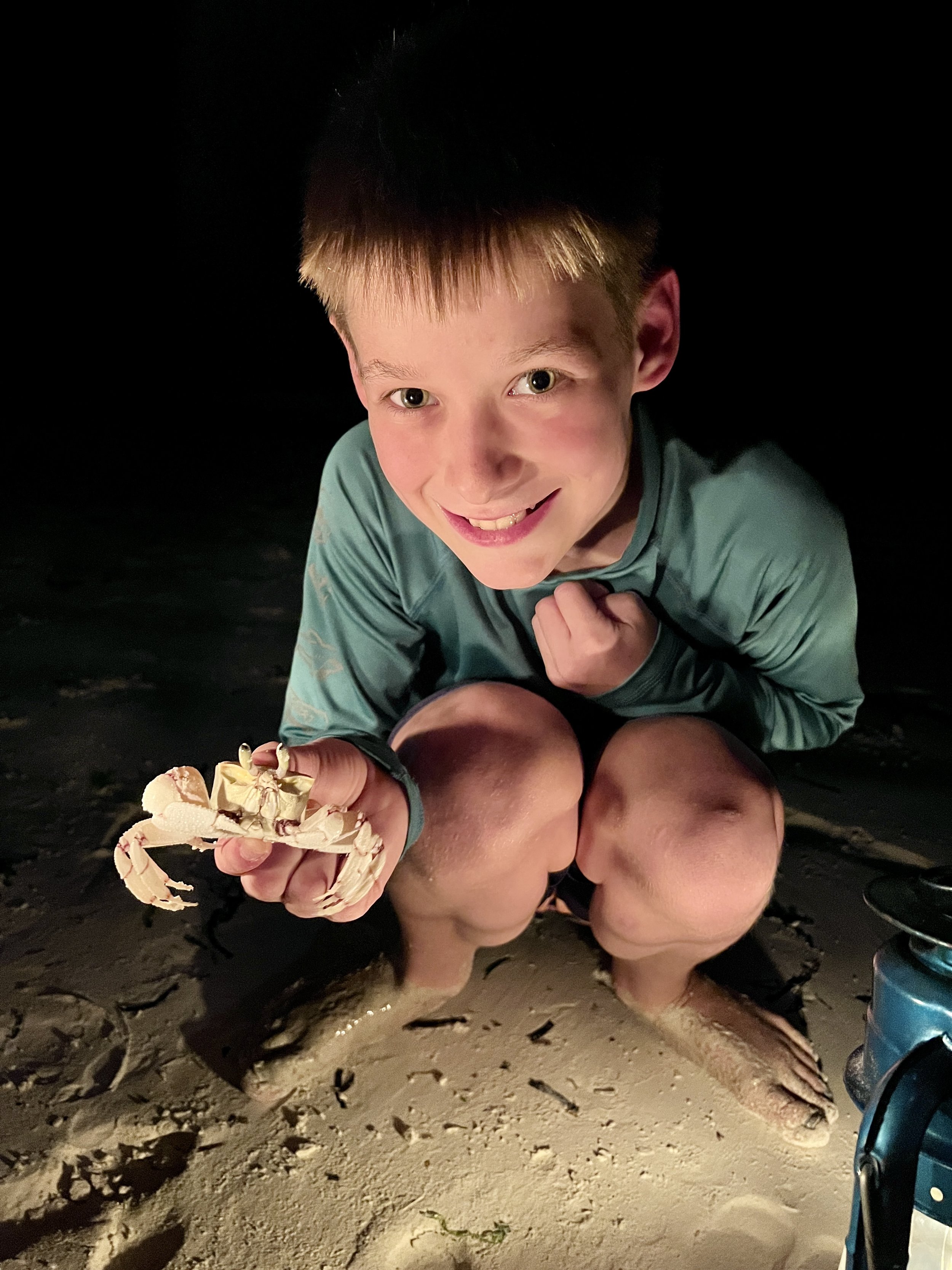 A young boy with blonde hair smiling and crouching on the sand at night, holding a small crab in his hand. The boy is wearing a teal long-sleeve shirt, and the scene is illuminated by a bright light, with a dark background.