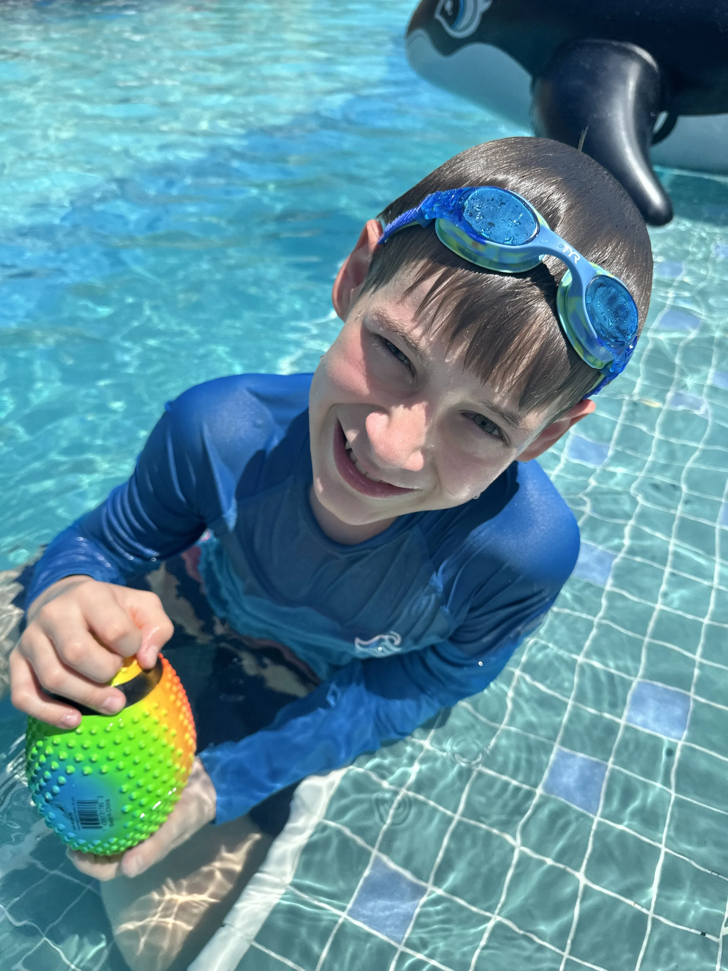 A young boy in a blue rash guard and swimming goggles smiling and holding a colorful textured ball in a swimming pool.