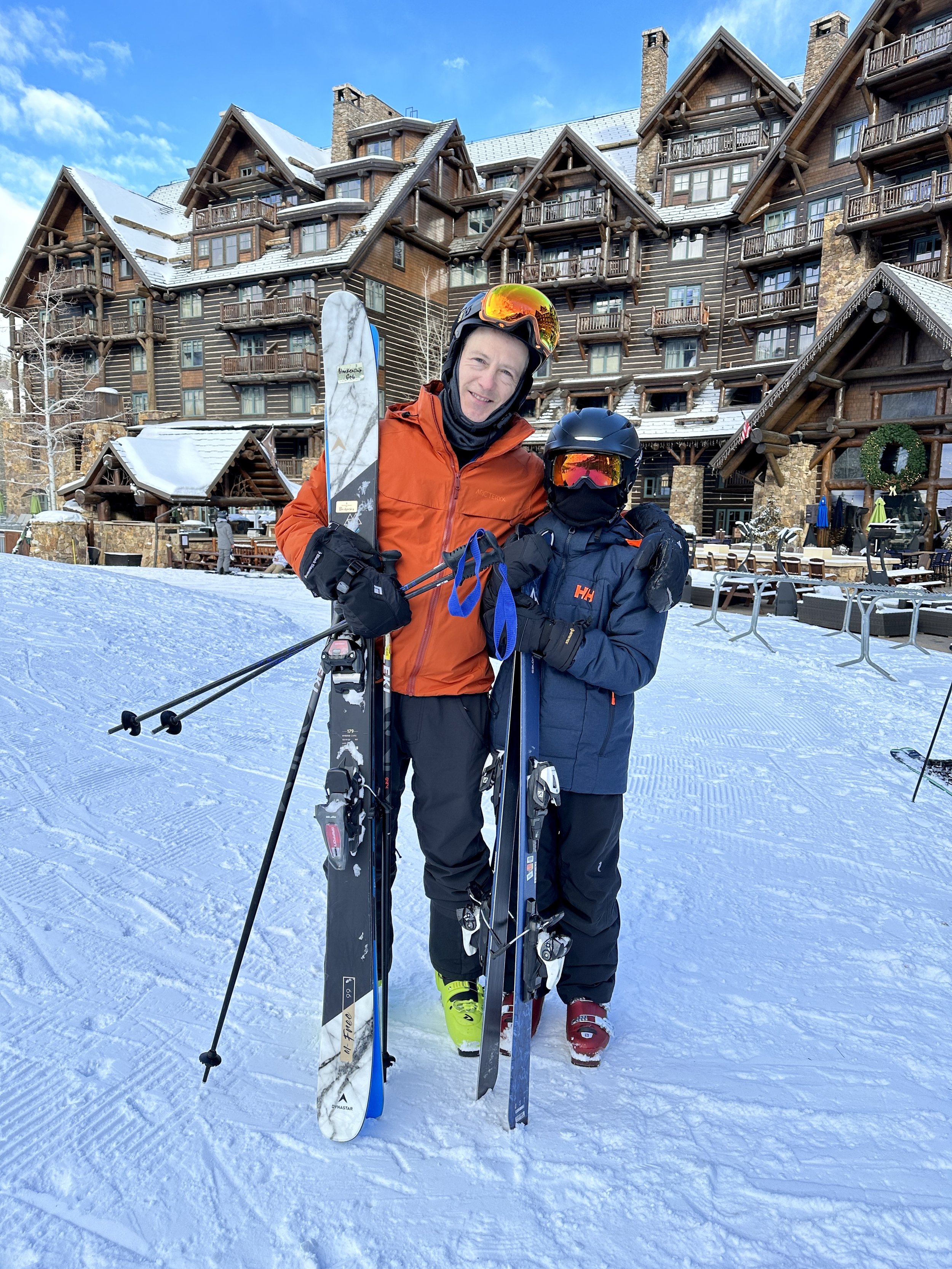Two skiers in winter gear standing on snow in front of a large wooden lodge with balconies, holding skis and ski poles, with snow-covered rooftops and blue sky in the background.