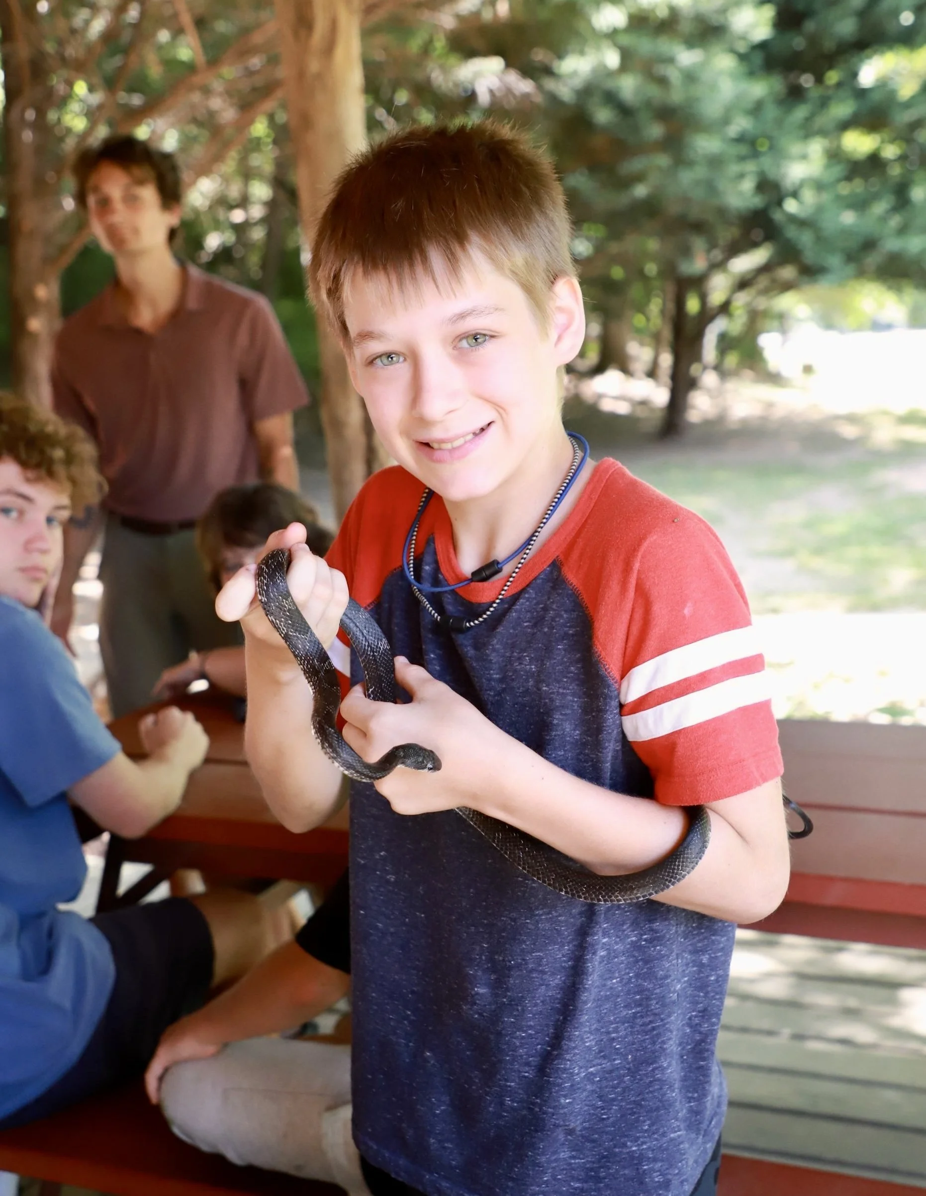 A smiling boy with short brown hair holding a black snake, with a group of boys and an adult in the background outdoors under trees.